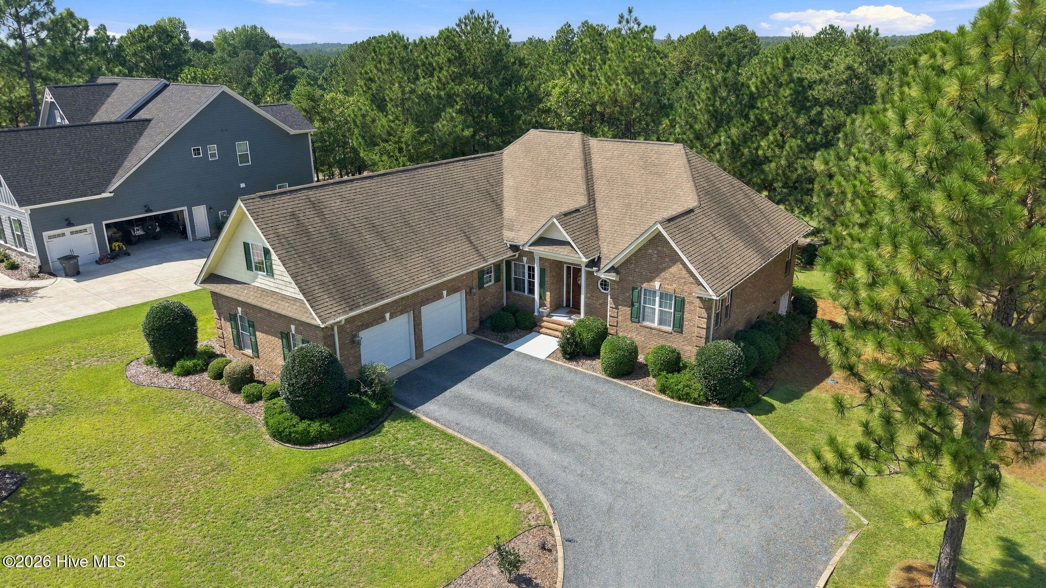 Aerial view of a brick home with a double garage, curved driveway, and pine trees.