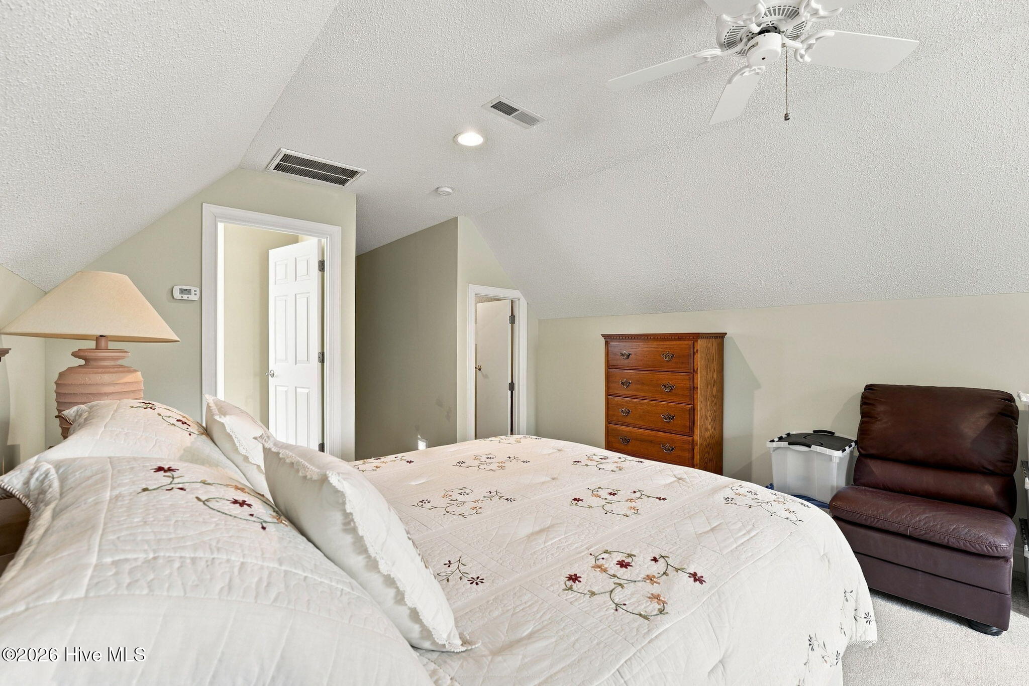 Bedroom with a floral quilted bed, wooden chest of drawers, and brown recliner beneath a sloped ceiling.