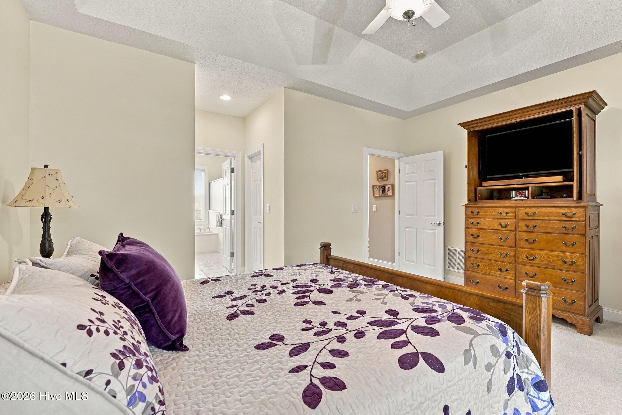 Bedroom with a purple-and-gray leaf-patterned quilt, tray ceiling, and tall wooden dresser with a television.