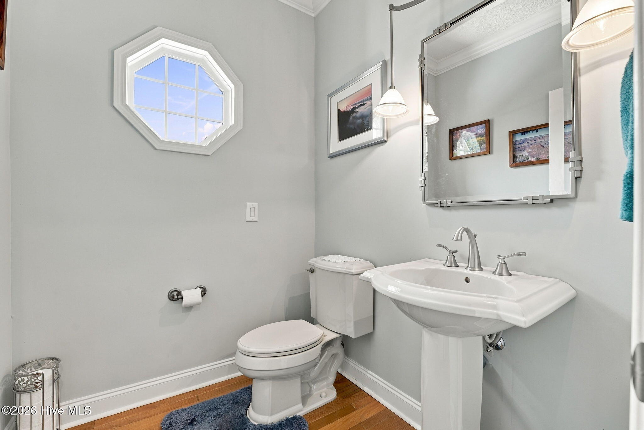 Bathroom with pedestal sink, toilet, and an octagonal window above hardwood flooring.