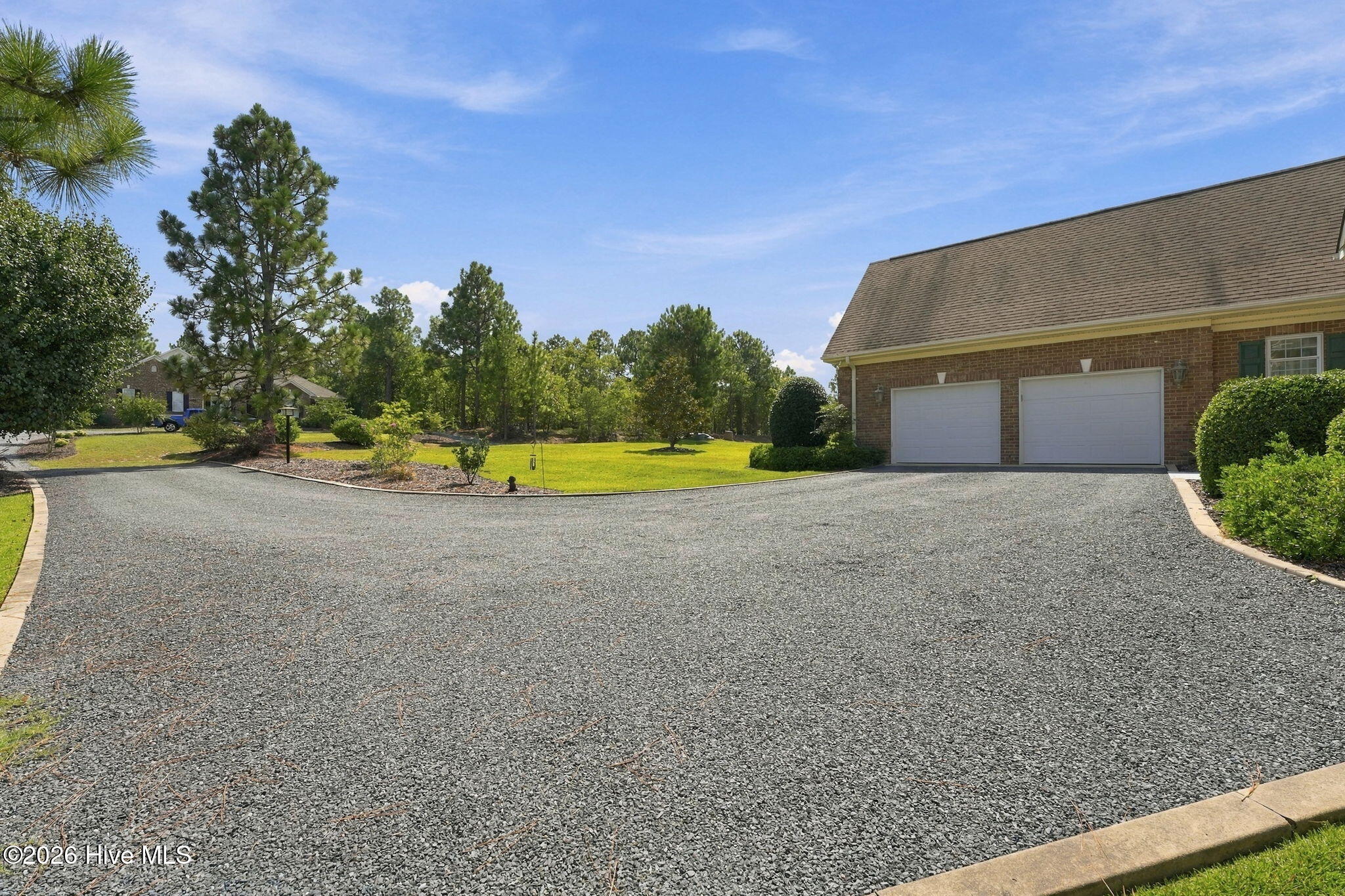 Gravel driveway leading to a brick house with two garage doors, bordered by a lawn and trees.