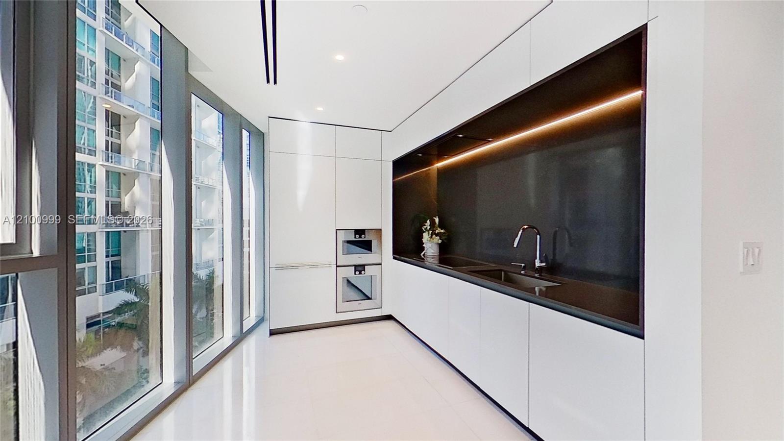 Modern white kitchen with dark stone backsplash, built-in double ovens, and floor-to-ceiling windows.