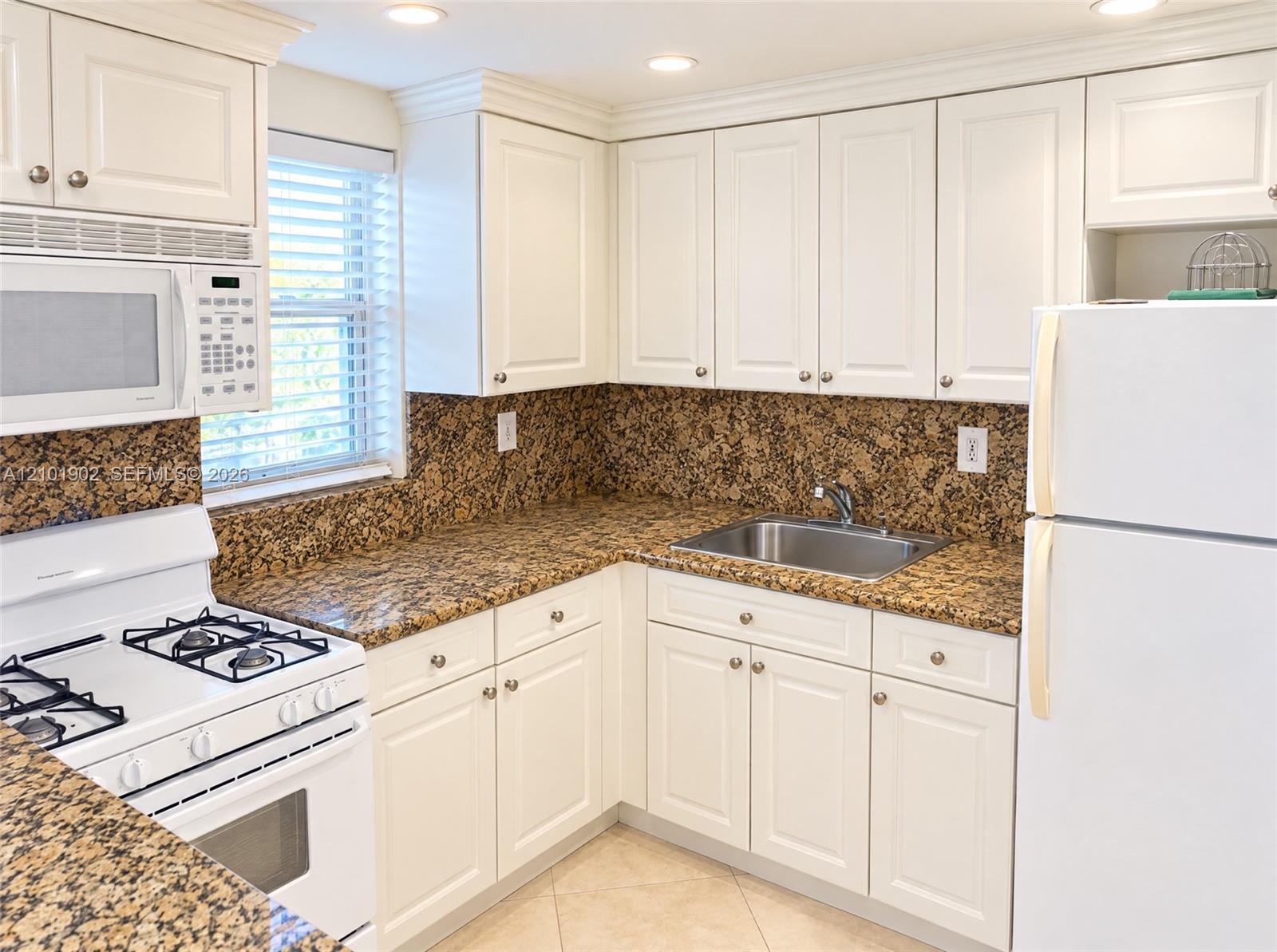 Condo kitchen with white cabinets, speckled granite counters, gas range, and window blinds.