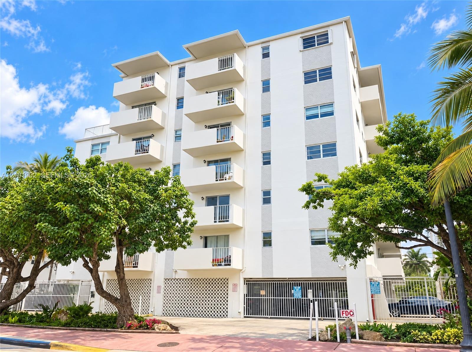White condominium building with projecting balconies, tropical trees, and a gated parking entrance.