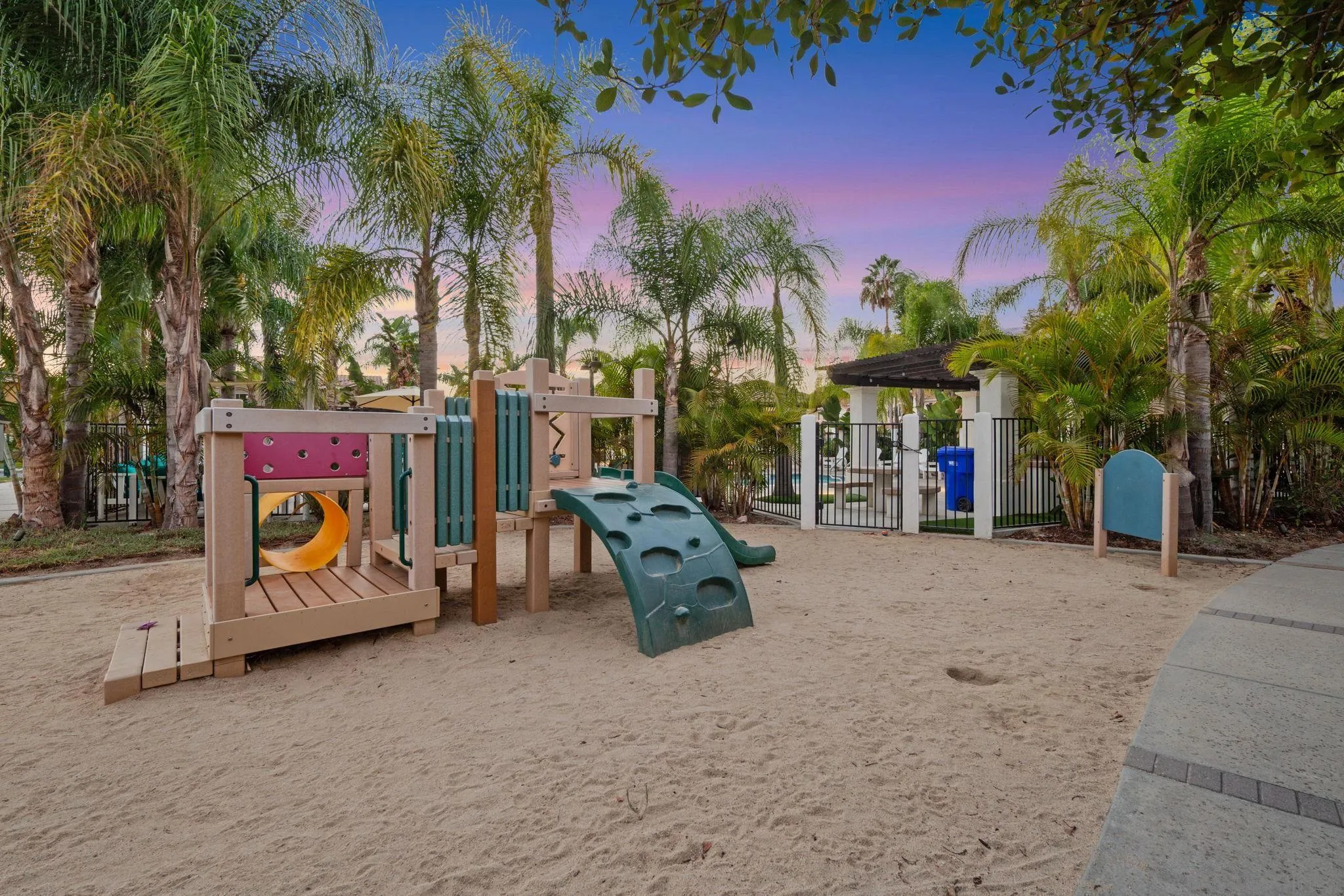 Sandy playground with a climbing structure and slide surrounded by palm trees at dusk.