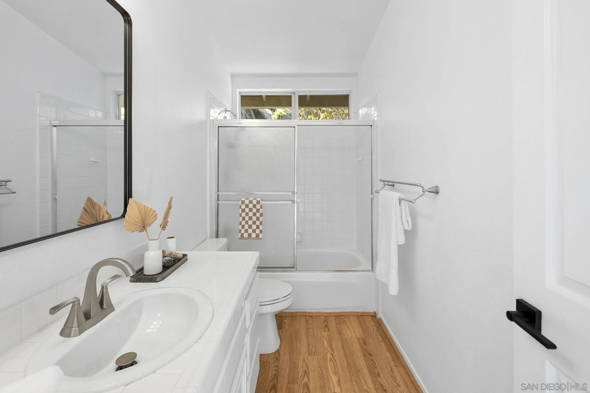 Bright bathroom with a white vanity, glass-enclosed tub and shower, and warm wood flooring.