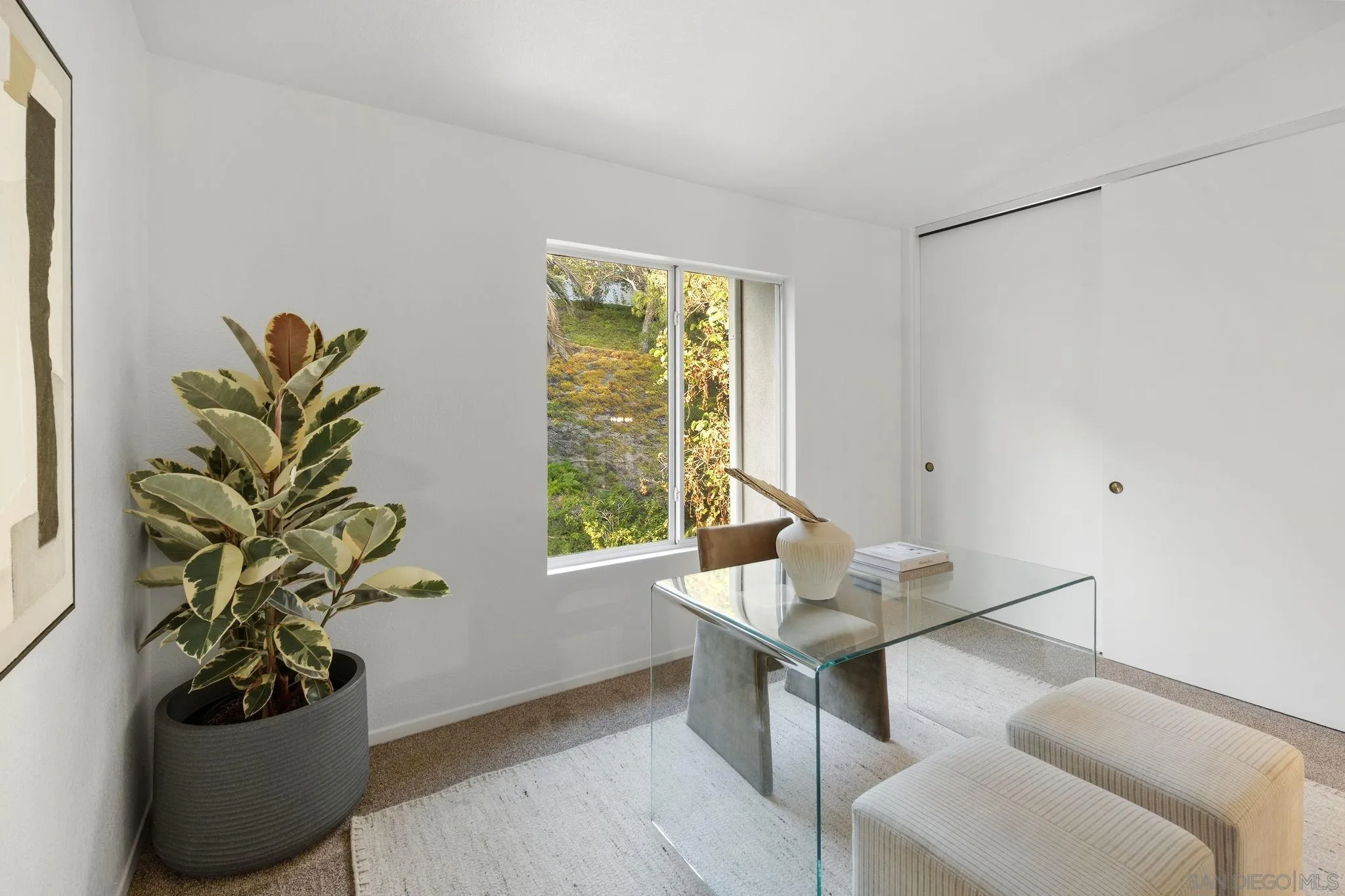 Bright home office with a glass desk, upholstered stools, and a window overlooking greenery.