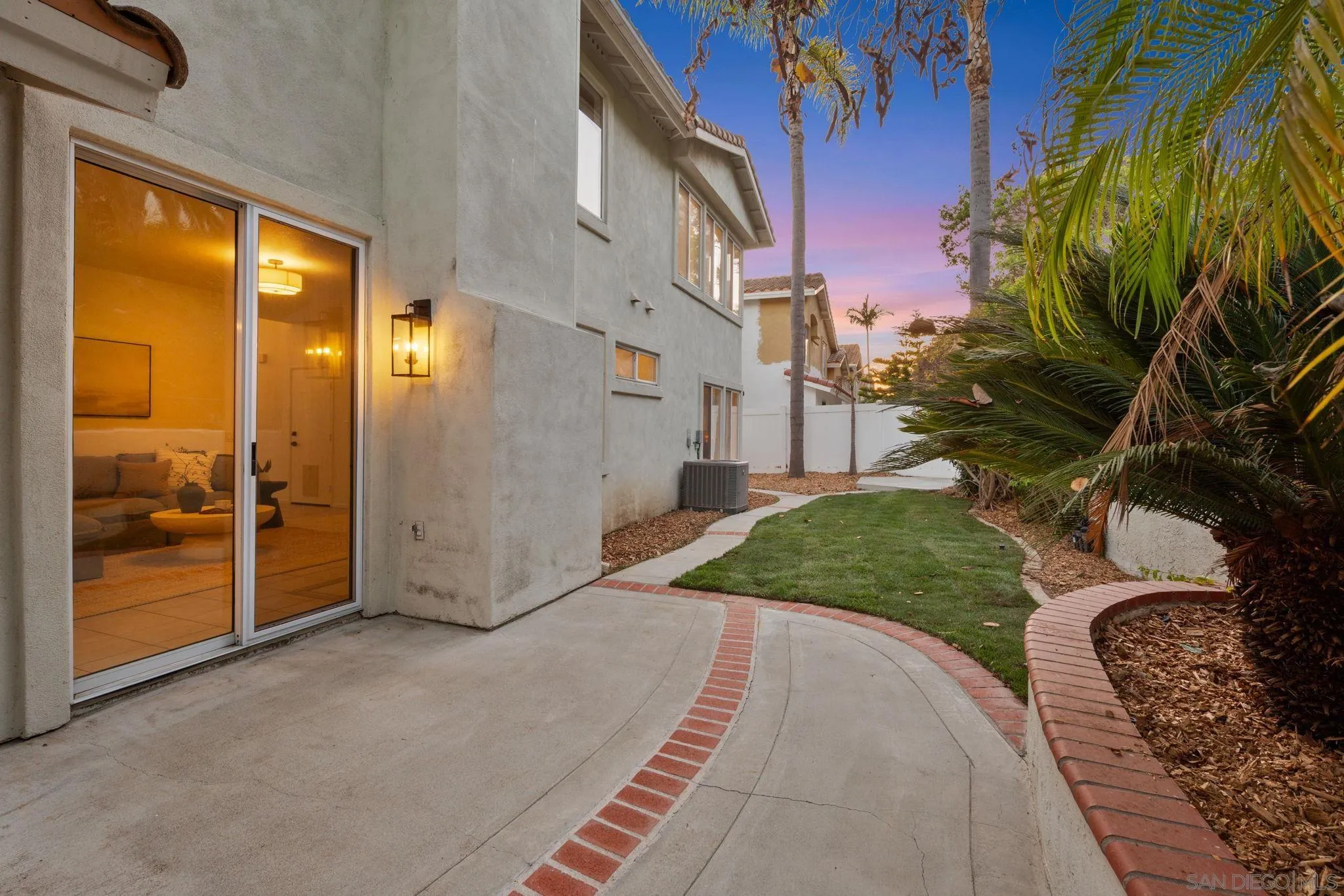 Home side yard with a curved brick-edged walkway, lawn, palms, and a warmly lit sliding glass door at dusk.