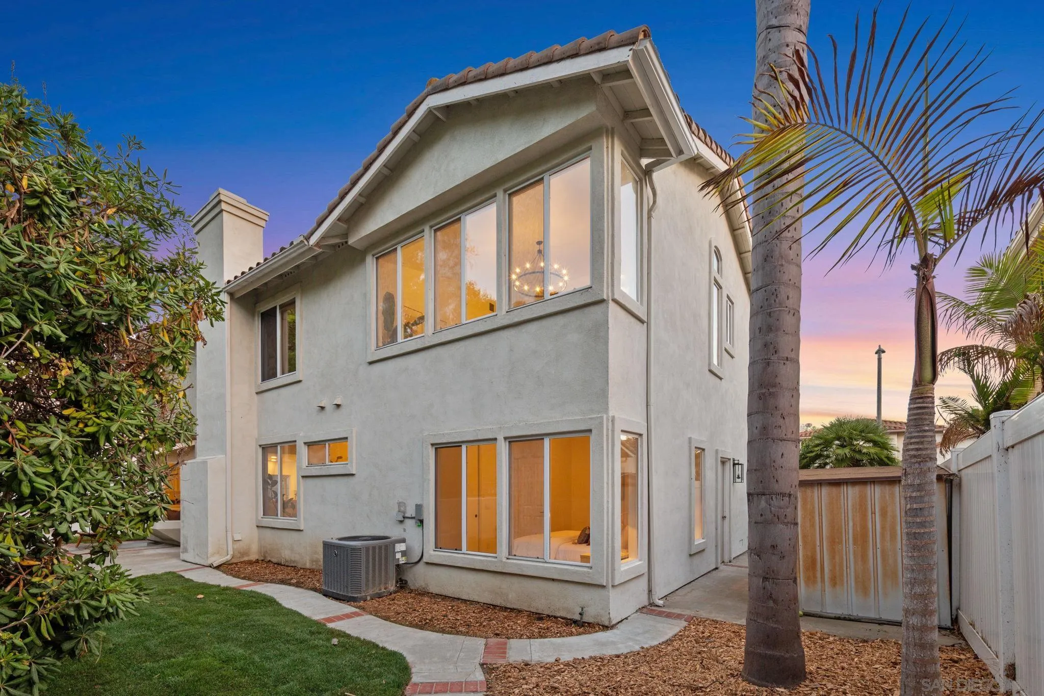 Two-story stucco home at dusk, with warmly lit windows and palm trees beside a small lawn.