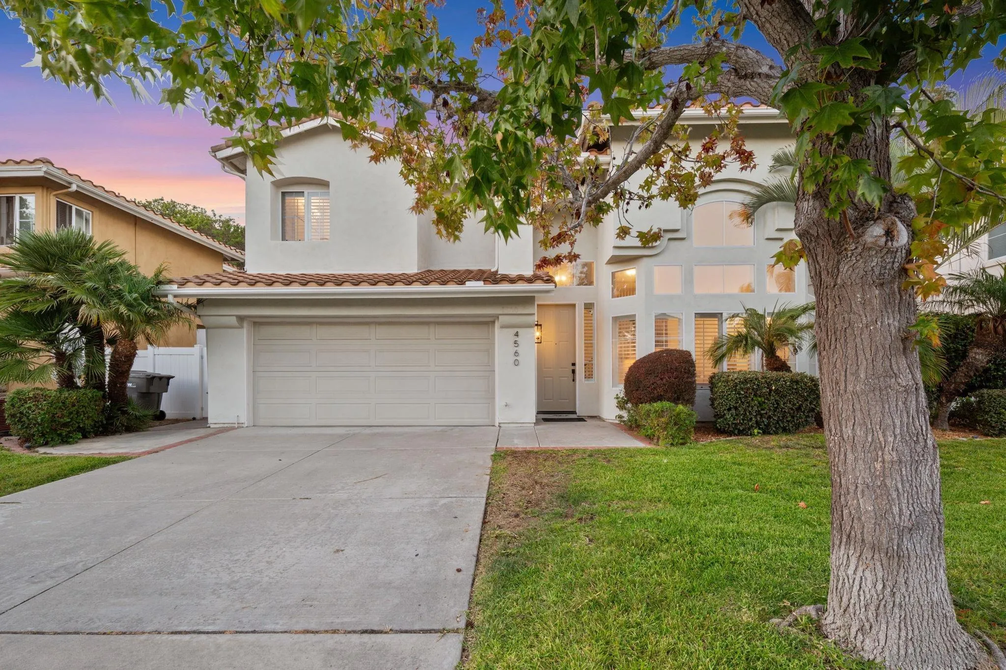 Two-story white home with a tiled roof and double garage, framed by a large tree at dusk.