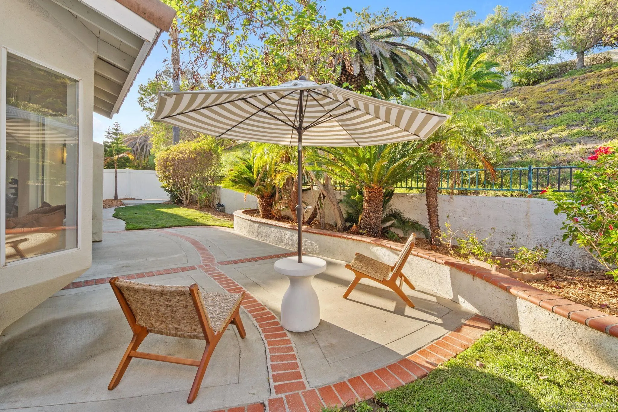 Outdoor patio with a striped umbrella, woven chairs, and tropical landscaping.