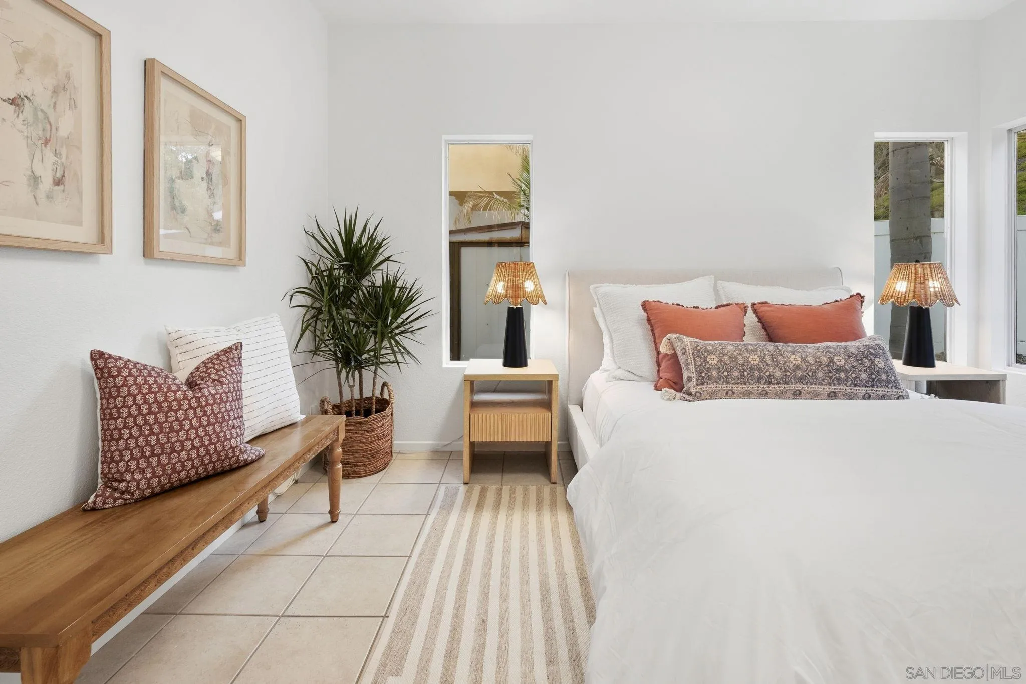 Bright staged bedroom with a white bed, rust-colored pillows, woven lamps, and a wooden bench.