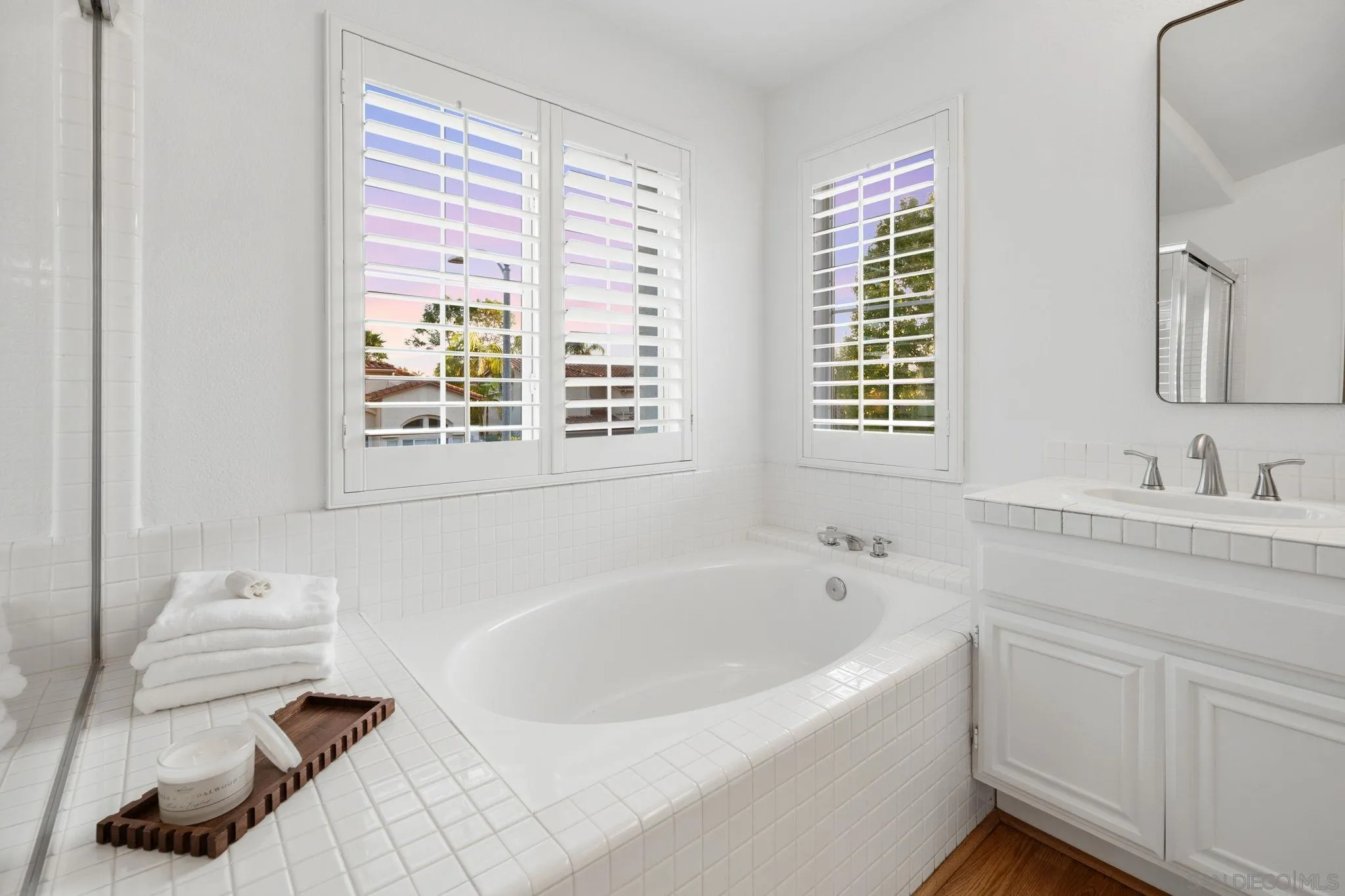 White tiled bathroom with a built-in soaking tub, shuttered windows, and a white vanity.
