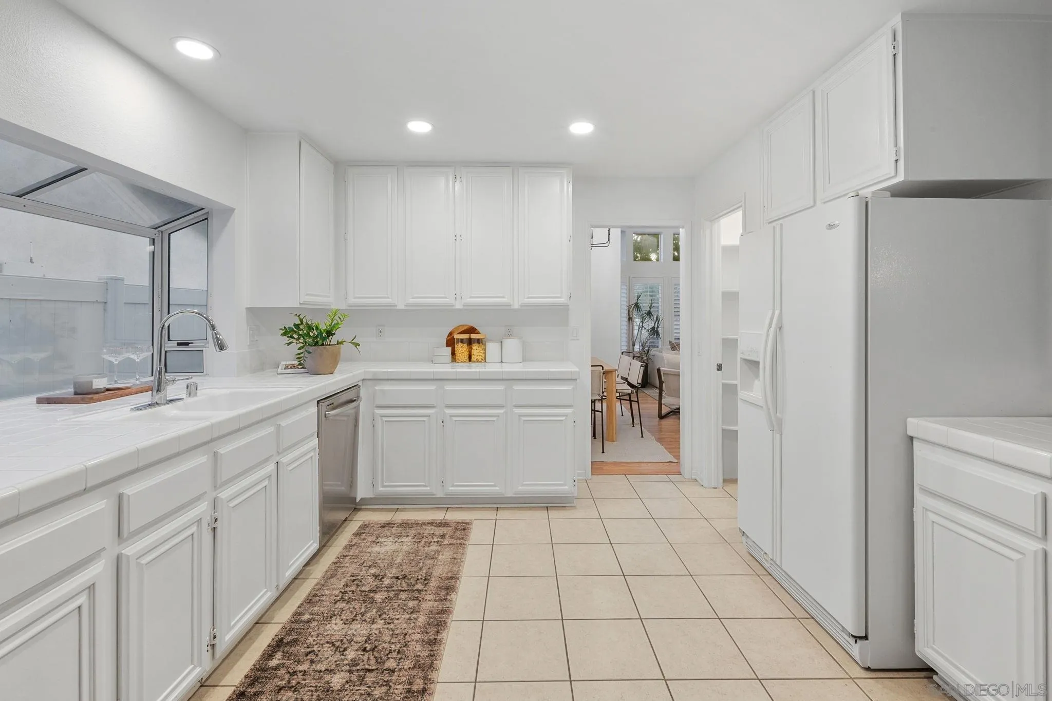 White kitchen with tiled counters and floor, a large refrigerator, and a doorway to the dining area.