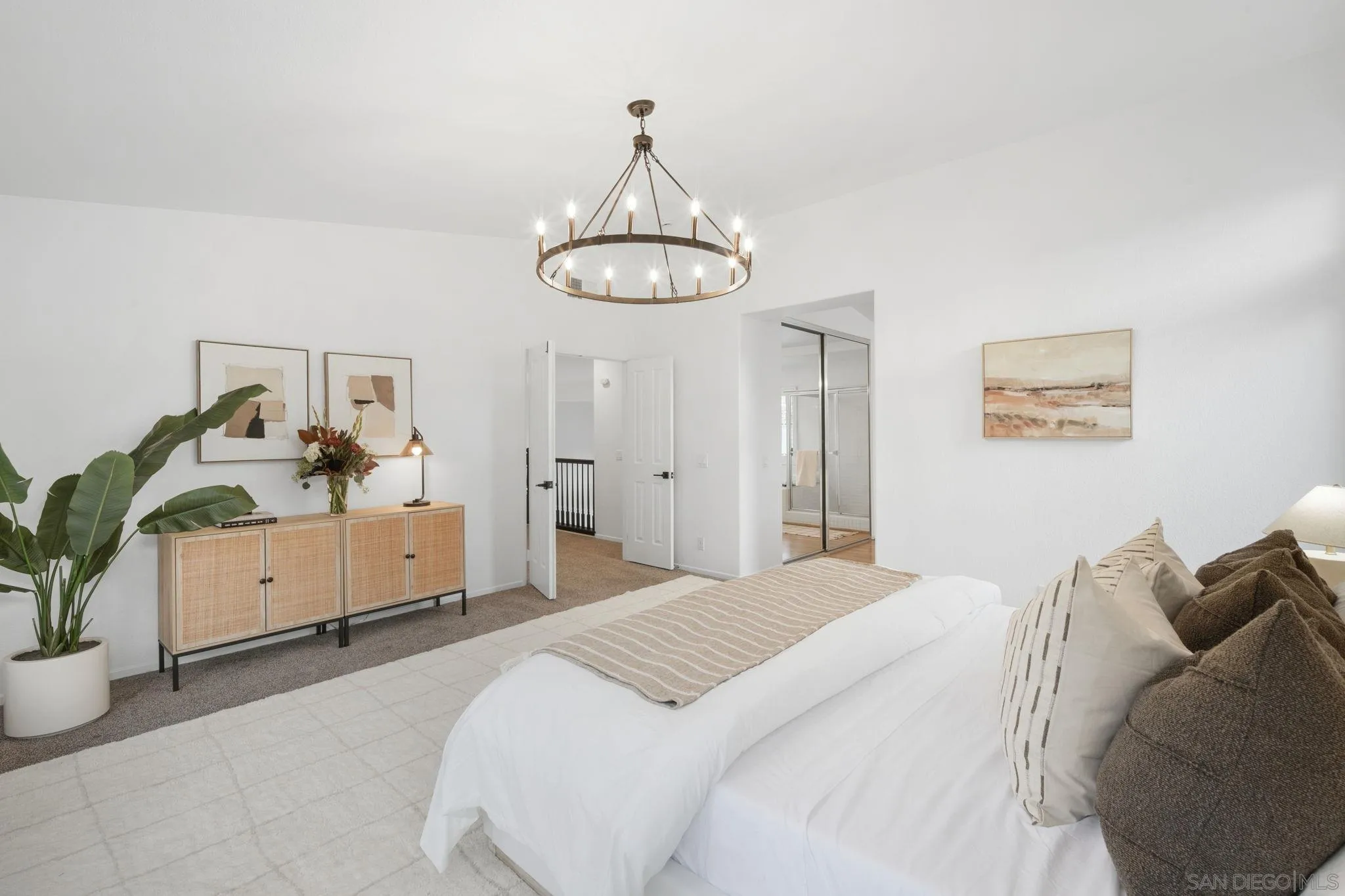 Staged bedroom with a white bed, brass ring chandelier, cane-front cabinet, and mirrored closet.