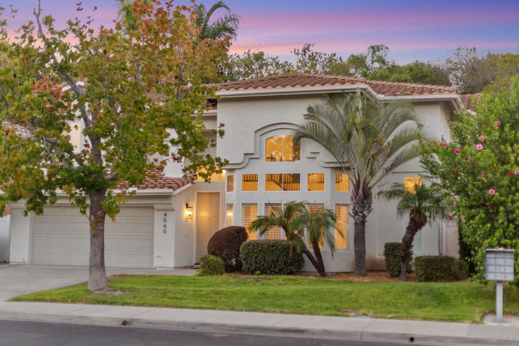 Two-story stucco home with a tile roof, palm trees, and warmly lit windows at dusk.