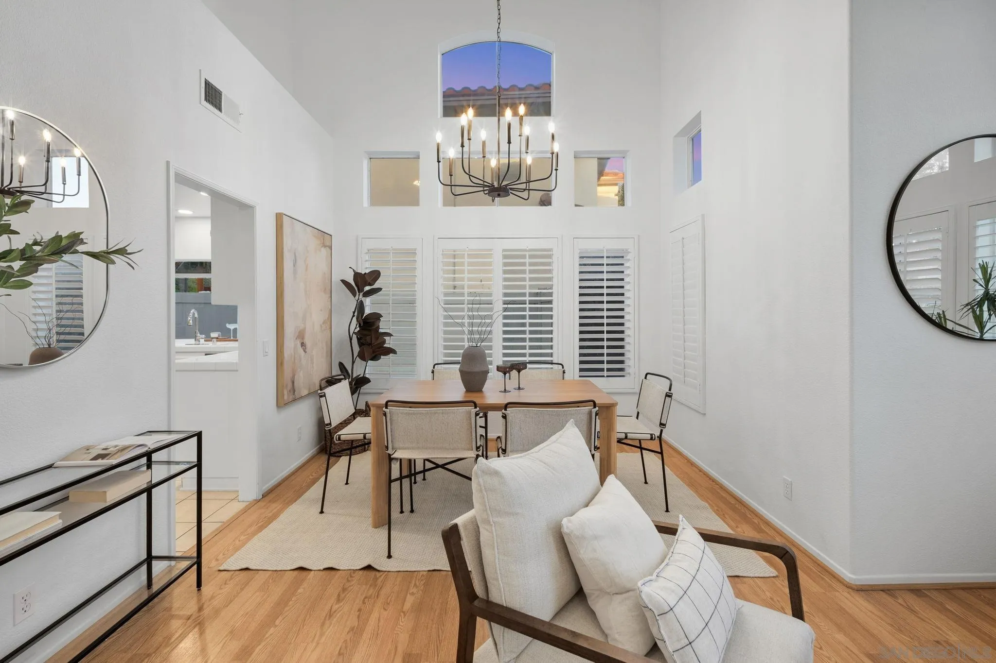 Dining room with vaulted ceiling, black chandelier, plantation shutters, and a wood table.