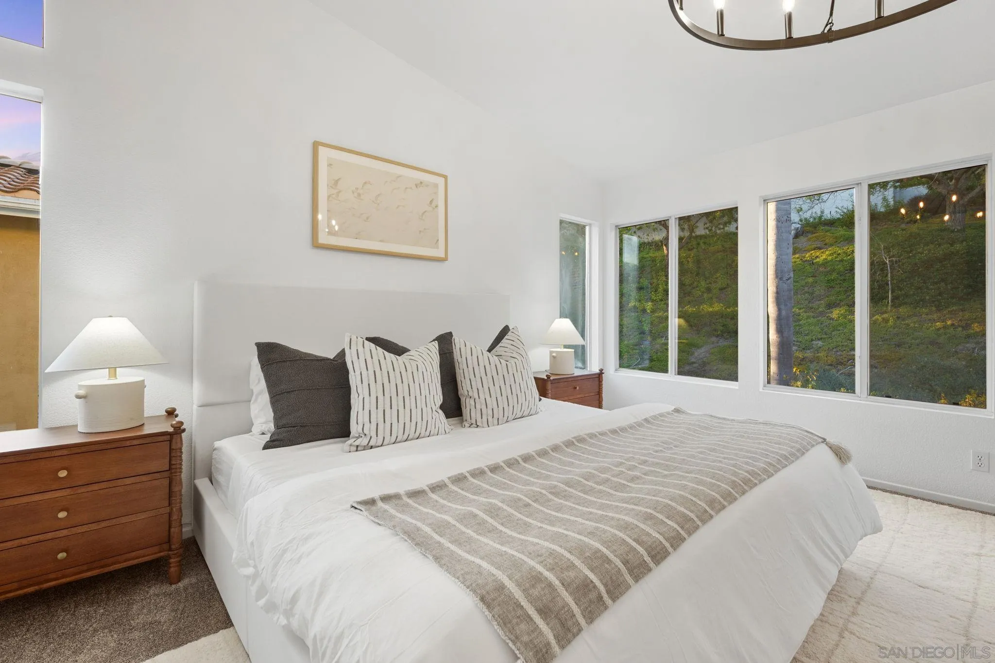 Bedroom with a white bed, striped throw, wooden nightstands, and windows overlooking a green hillside.