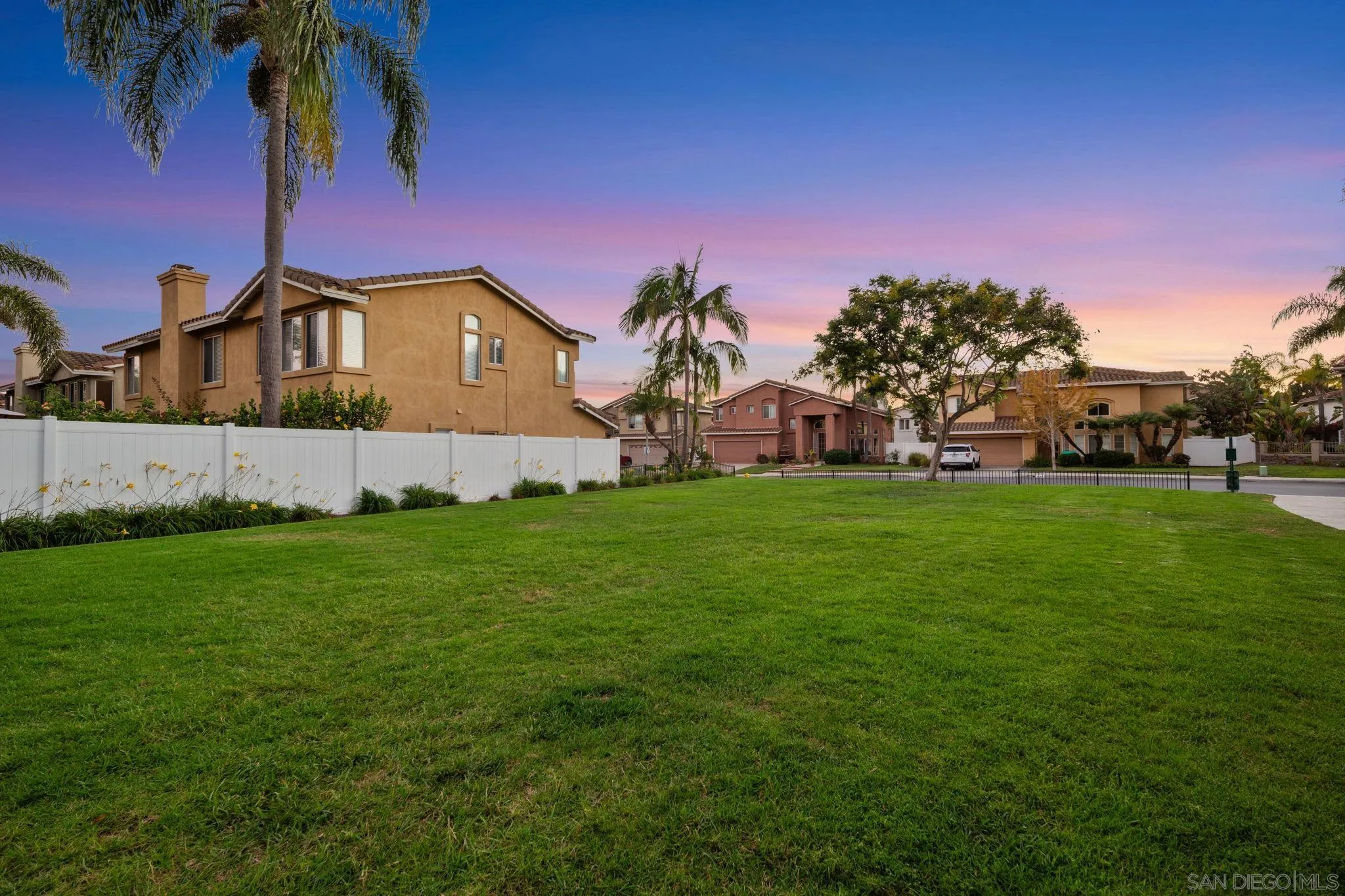Residential neighborhood with a broad green lawn, white fence, and palm trees at sunset.