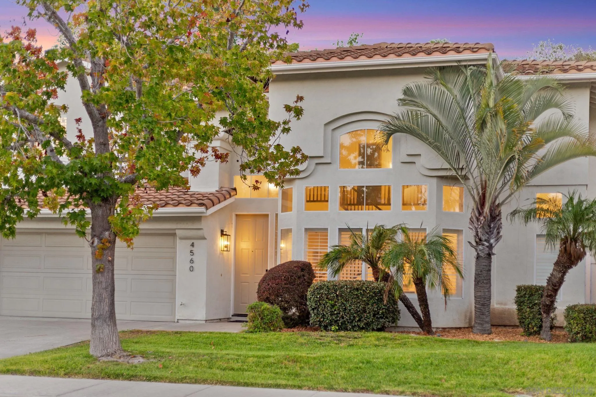 Two-story stucco home with a Spanish tile roof, arched window, palm trees, and illuminated windows at dusk.