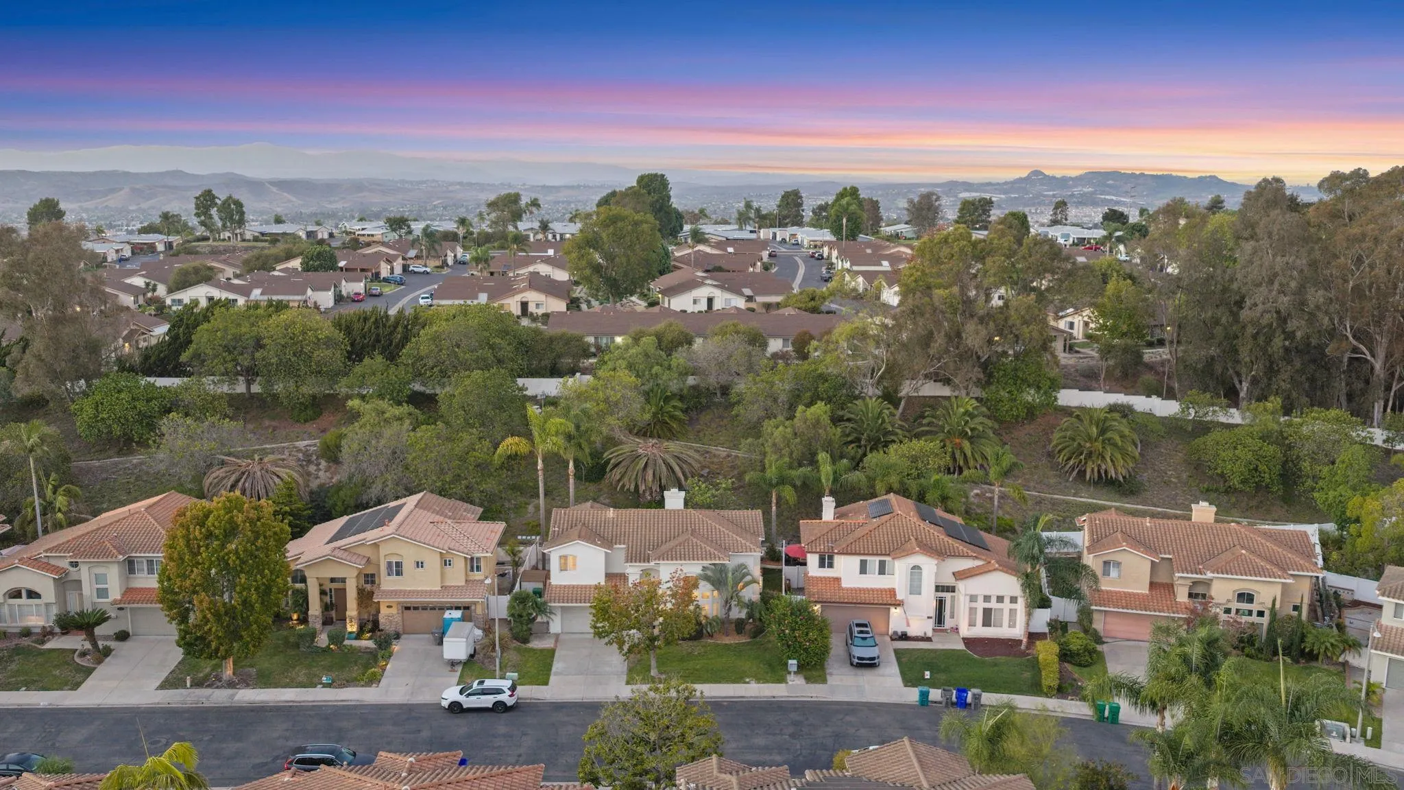 Aerial view of homes, landscaped yards, and distant hills beneath a colorful dusk sky.
