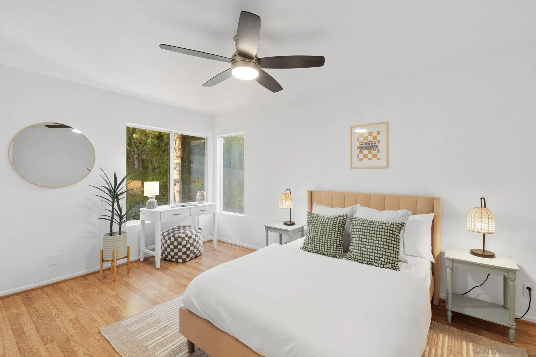Bedroom with a tan upholstered bed, patterned green pillows, wood floors, and large windows.