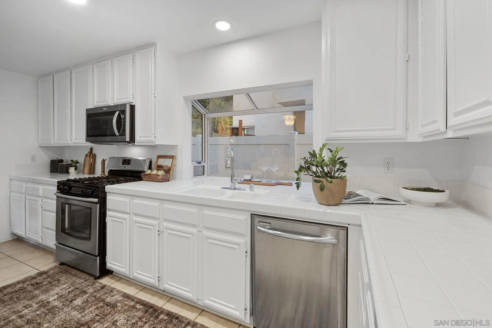 White kitchen with tiled counters, black range, stainless steel dishwasher, and a pass-through window.