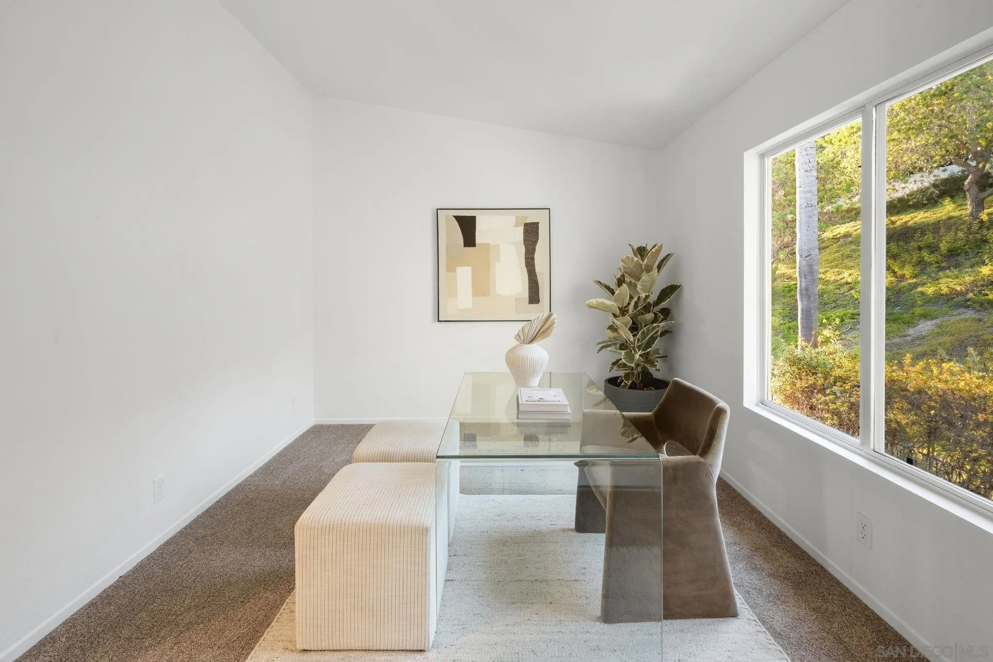 Dining room with a glass table and cream benches beside a large window overlooking greenery.