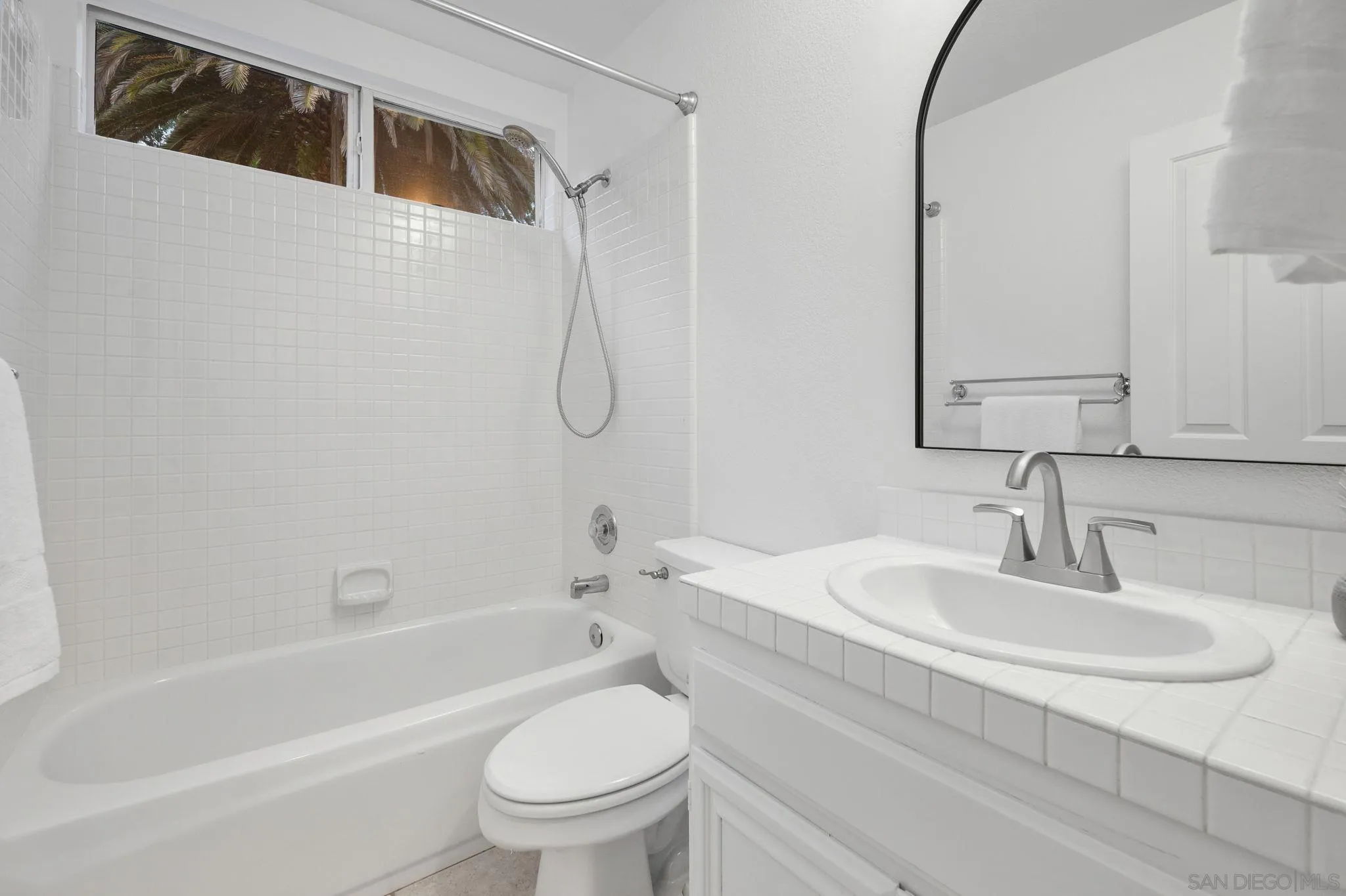 White bathroom with tiled tub and shower, high window, and tiled vanity with oval sink.
