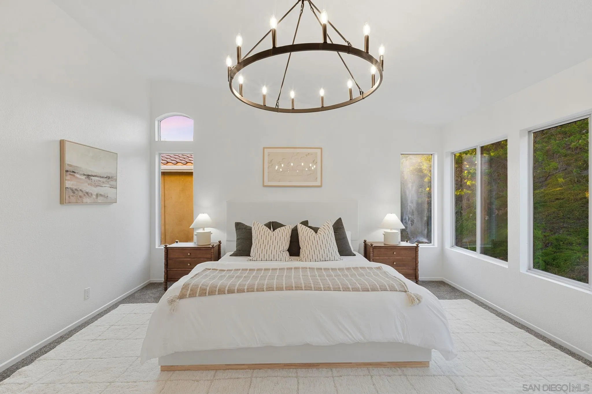 Bright staged bedroom with a centered bed, tall windows overlooking greenery, and a circular chandelier.