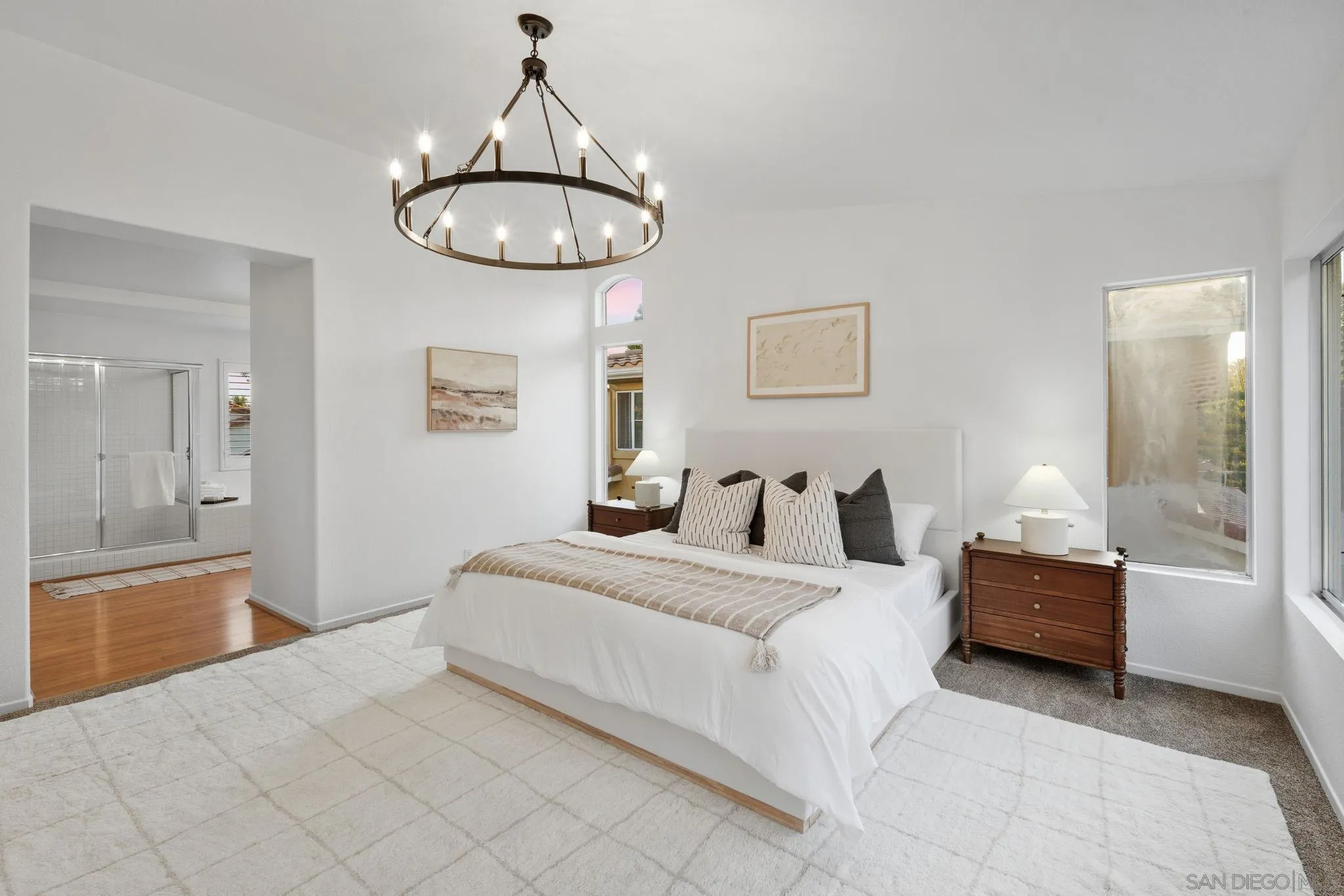 Staged bedroom with a white bed, circular chandelier, and adjoining glass-enclosed shower.