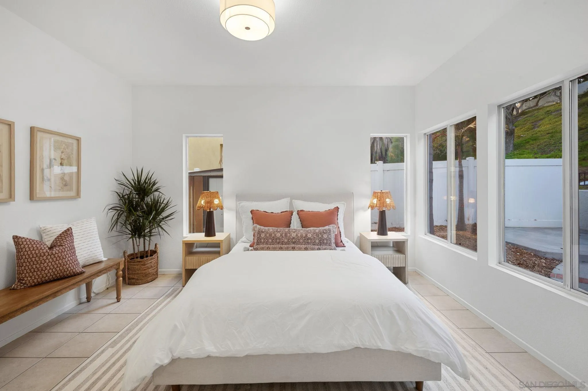 Bright staged bedroom with a white bed, rust-colored pillows, and large windows overlooking a fenced yard.