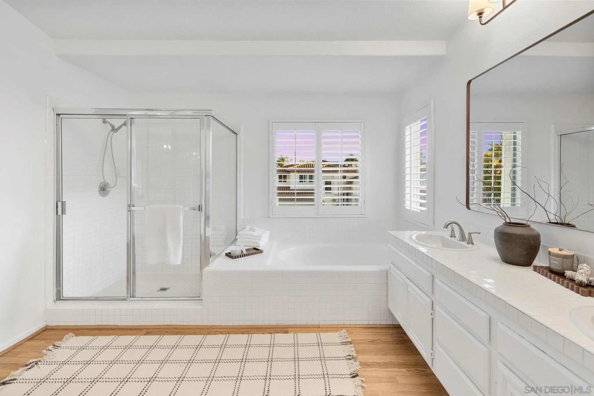 Bright bathroom with a glass shower, soaking tub, tiled vanity, and shuttered windows.