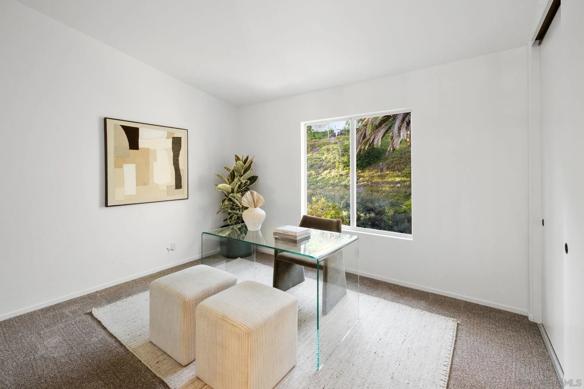 Staged home office with a glass desk, cream stools, and a window overlooking a green hillside.