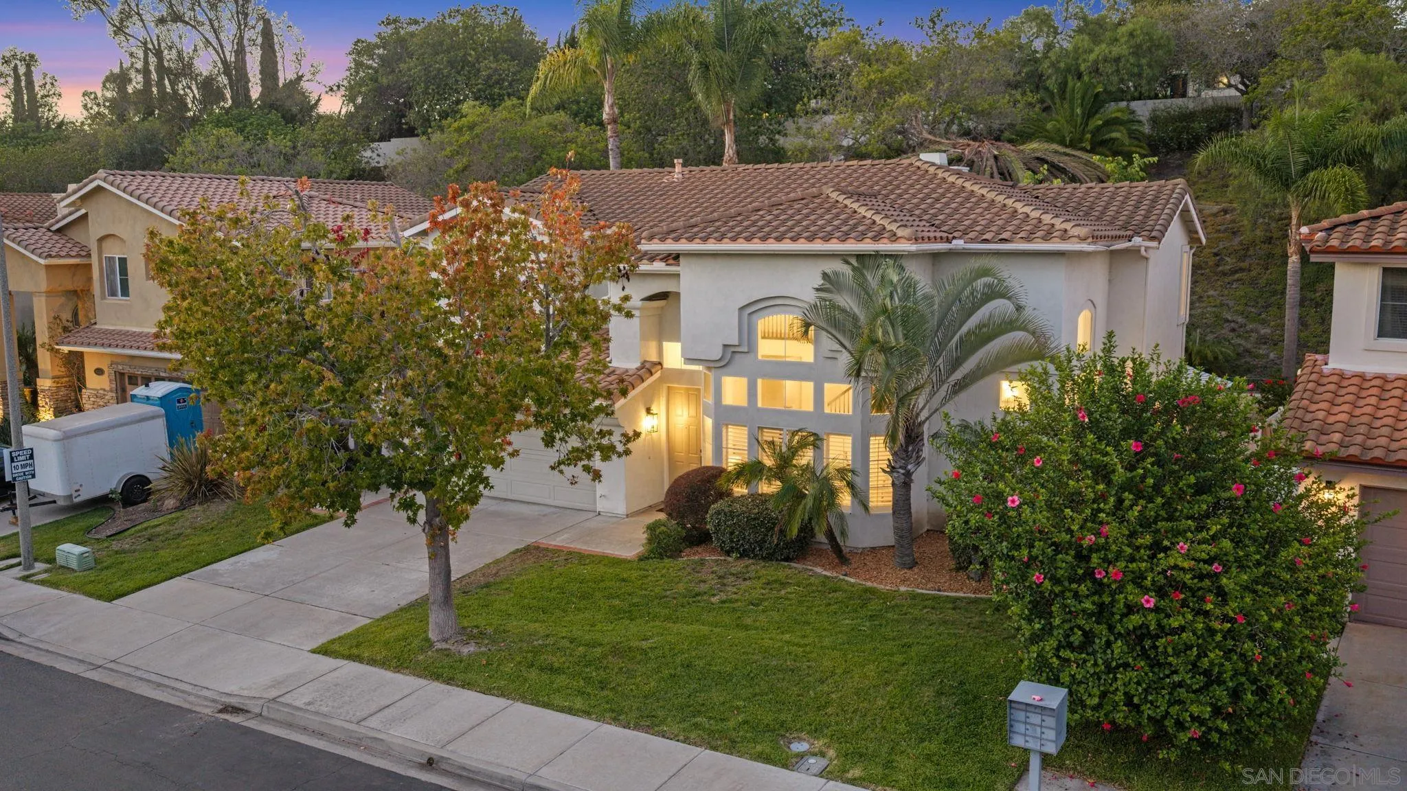 Two-story stucco home with a tiled roof, illuminated windows, and landscaped front yard at dusk.