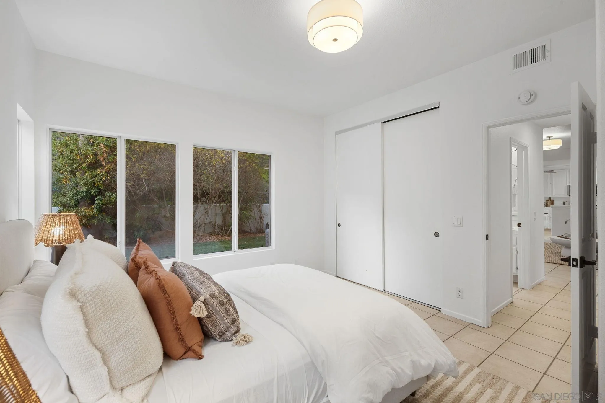 Bedroom with a white bed, broad windows overlooking a leafy fenced yard, and a sliding closet door.