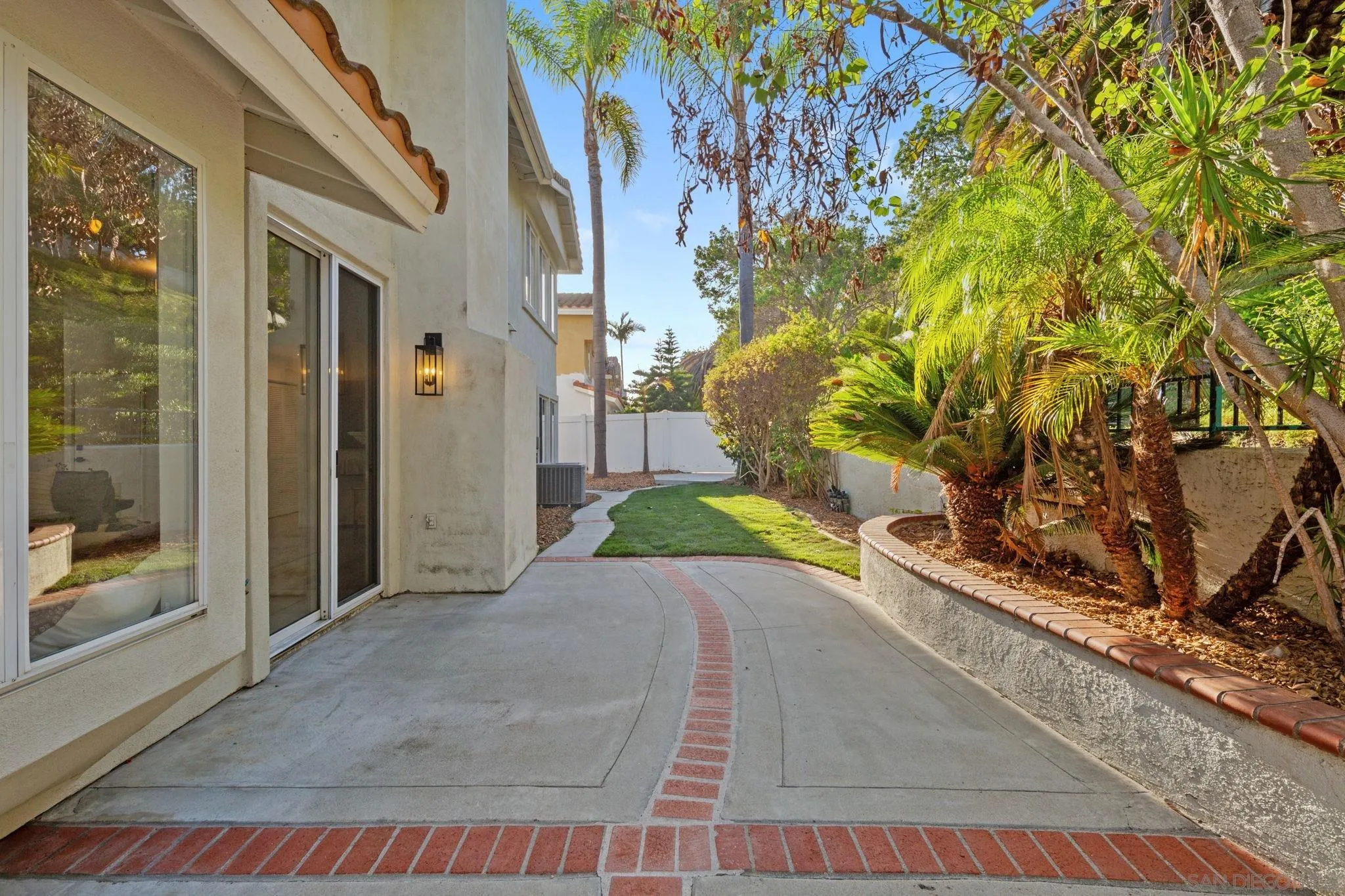 Curved concrete patio beside a stucco home, with brick inlay and a landscaped side yard with palm trees.
