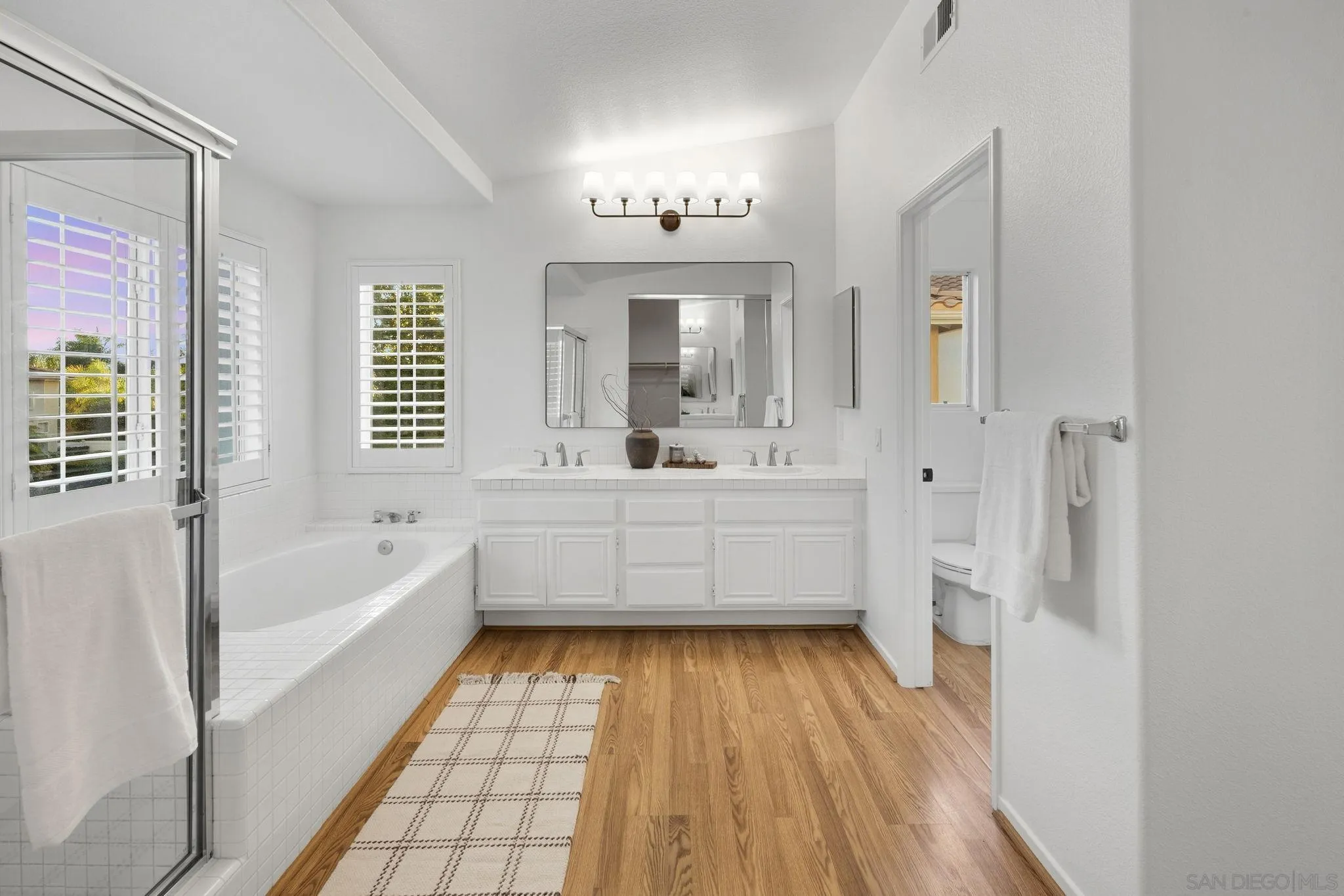 Bright bathroom with a double vanity, tiled soaking tub, glass shower, and warm wood flooring.