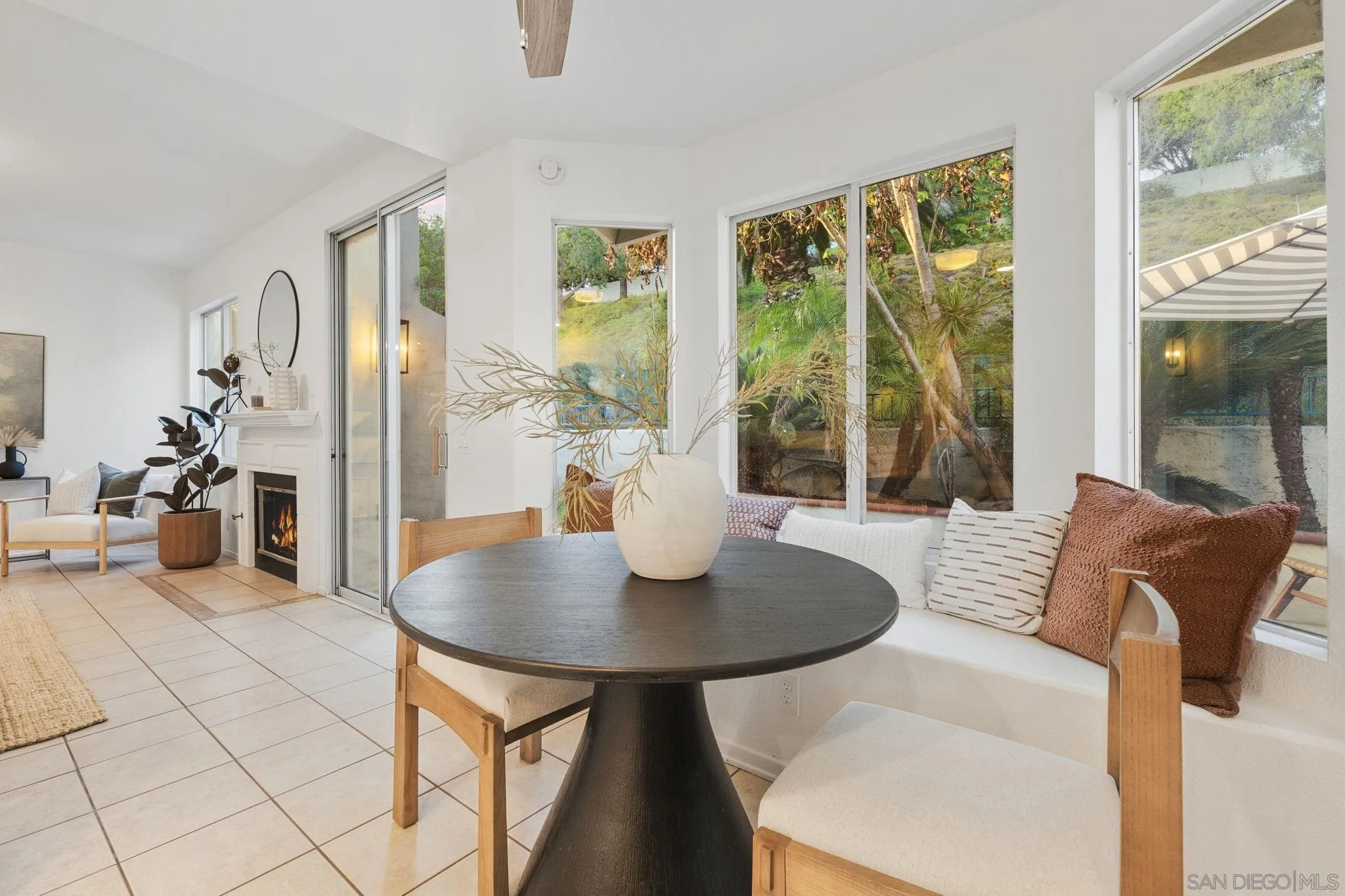 Dining area with round black table, cushioned window seating, and a fireplace beside large garden-facing windows.