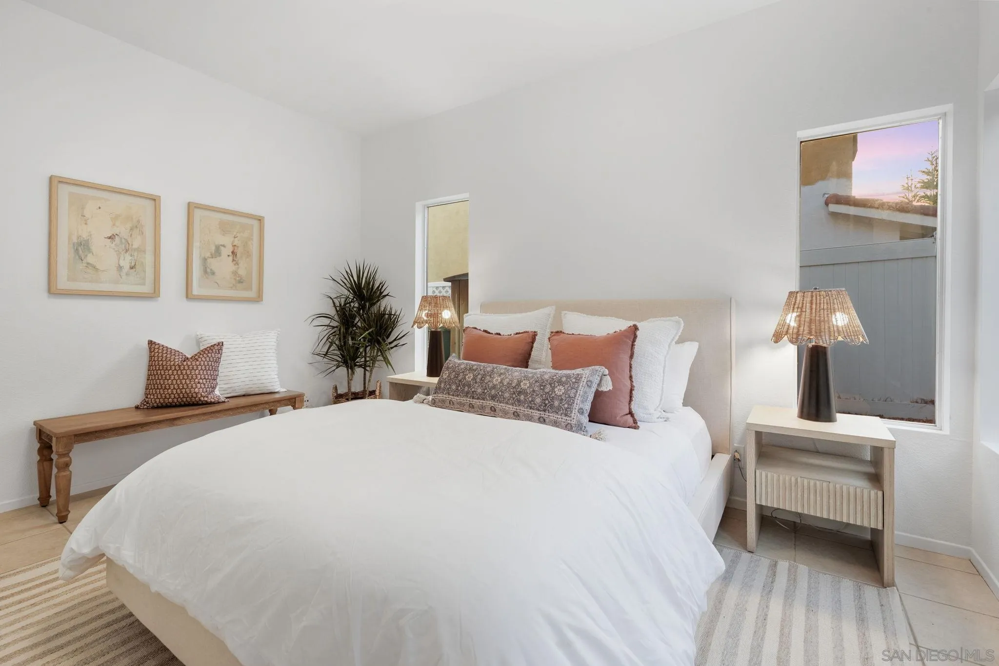 Staged bedroom with a white bed, rust-colored pillows, woven-shade lamps, and a wooden bench.