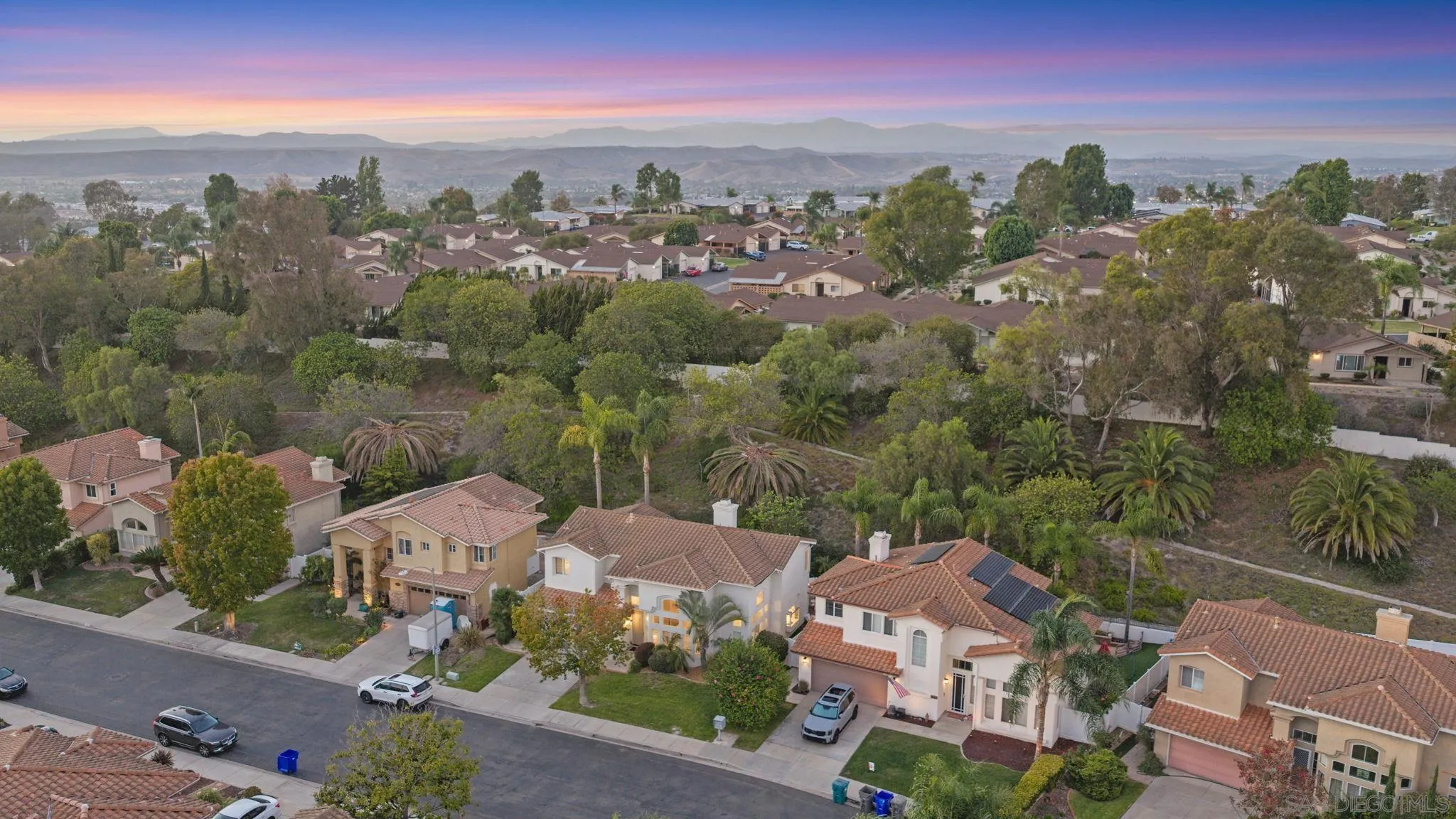 Aerial view of a residential neighborhood with palm trees and distant hills at sunset.