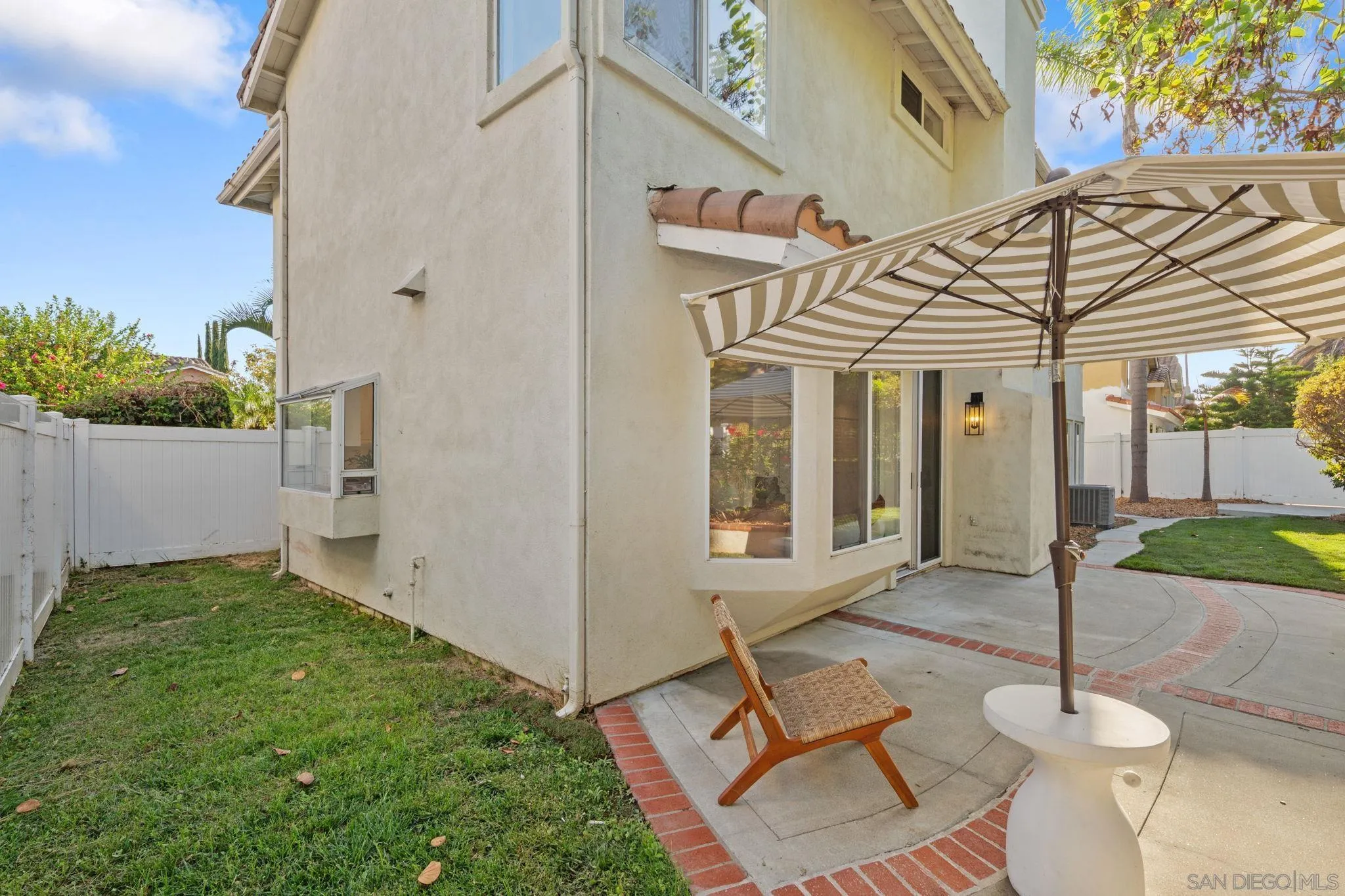 Home’s rear patio with a striped umbrella, curved paving, and fenced lawn.