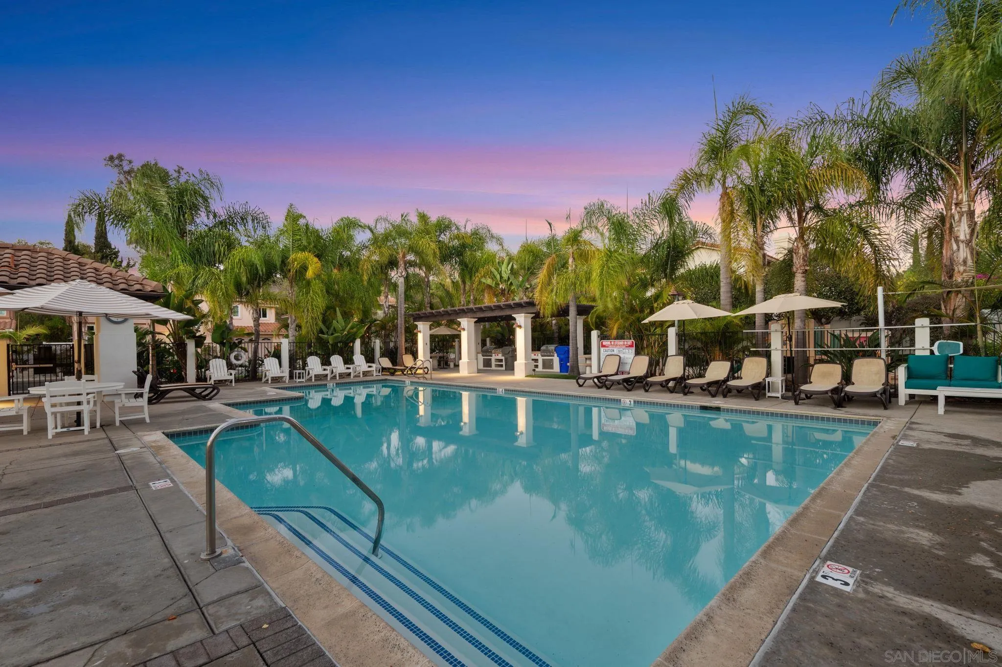 Outdoor pool with palm trees, lounge chairs, umbrellas, and a covered barbecue area at dusk.