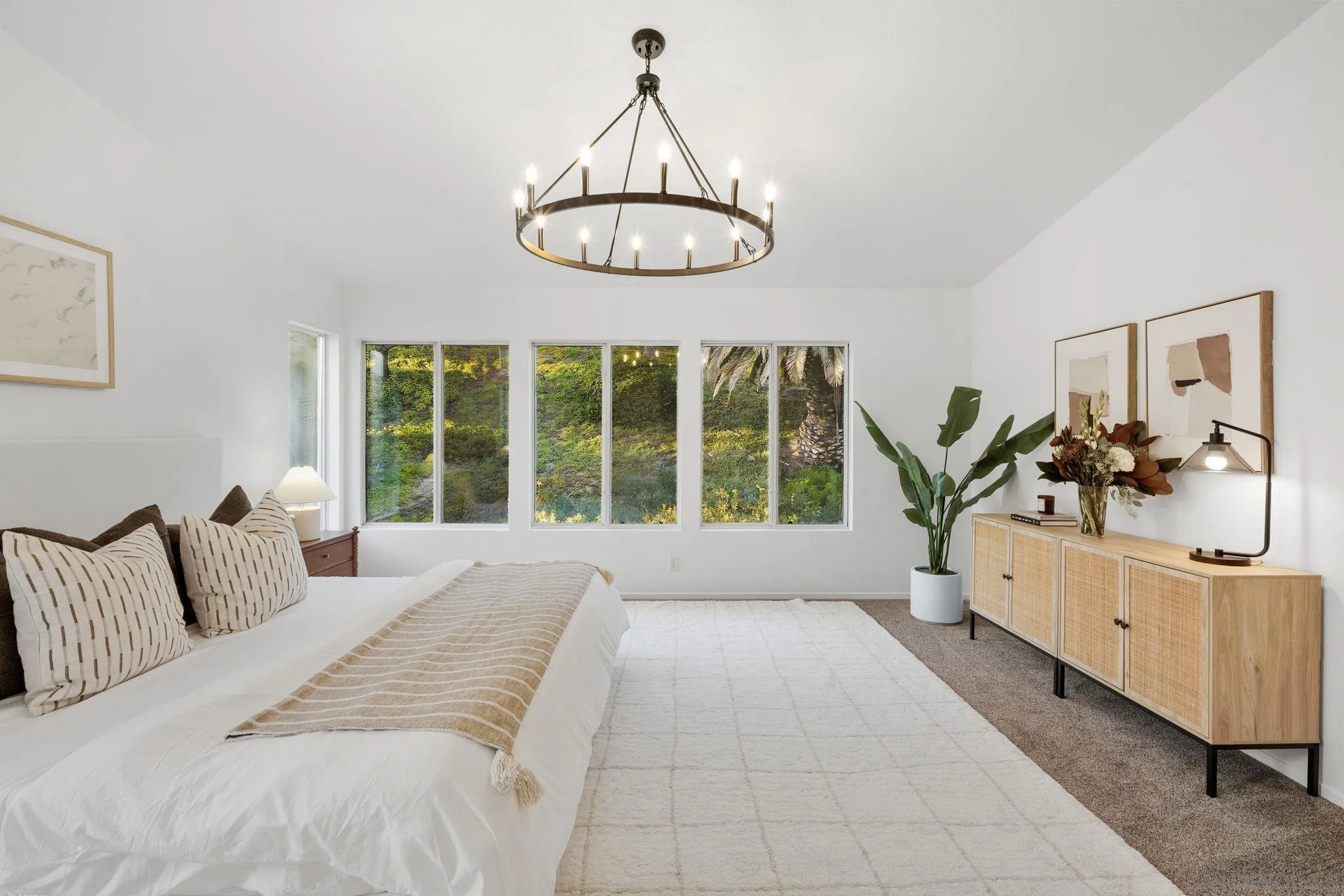 Staged bedroom with a white bed, brass ring chandelier, and large windows overlooking greenery.
