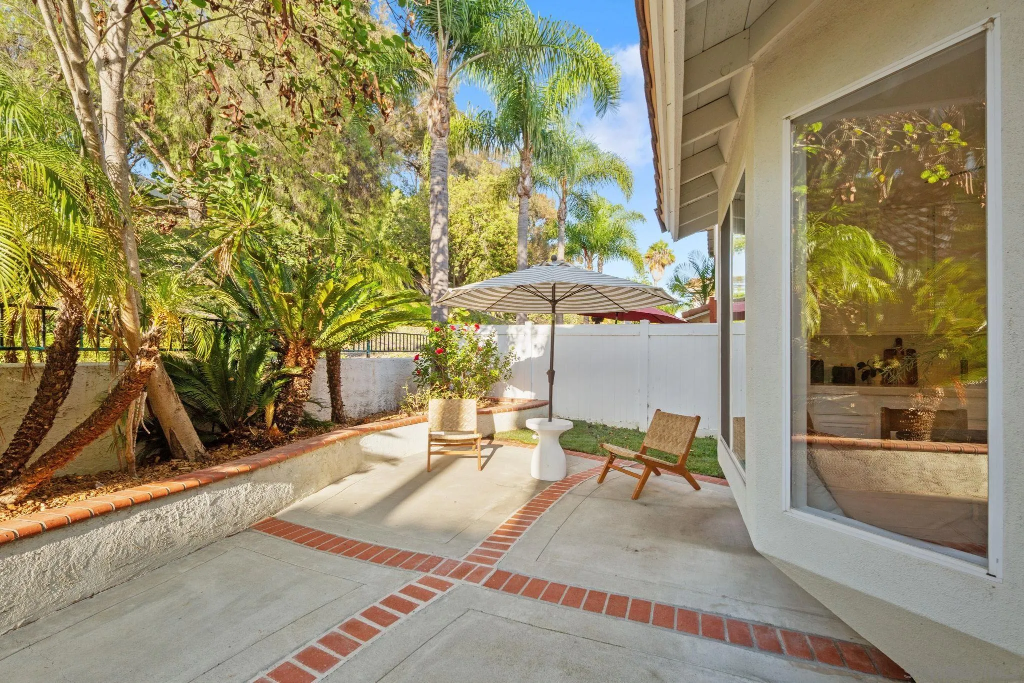 Backyard patio with striped umbrella, lounge chairs, brick edging, and lush tropical landscaping.