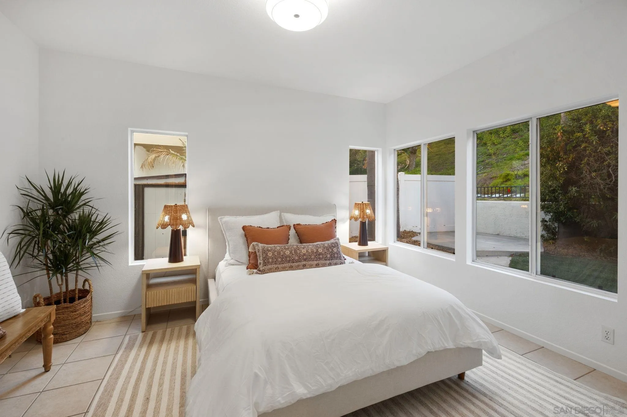 Staged bedroom with a white bed, rust-colored pillows, and large windows overlooking greenery.