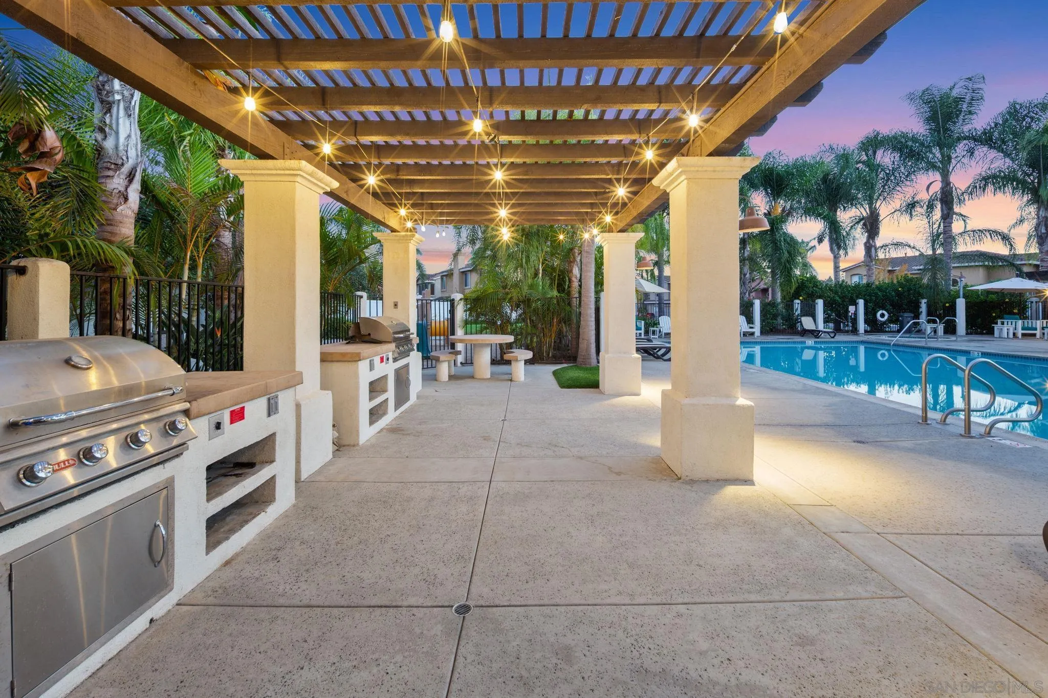 Outdoor poolside barbecue area beneath a lit wooden pergola, with stainless-steel grills and palm trees.