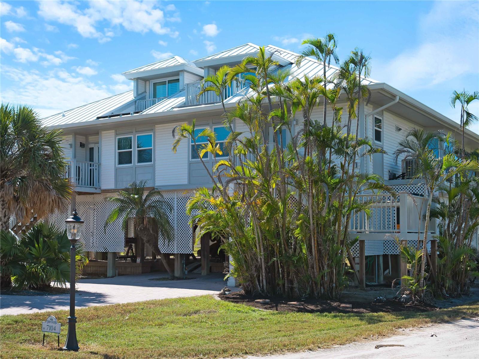 Elevated white coastal house with a metal roof and palm trees around it.