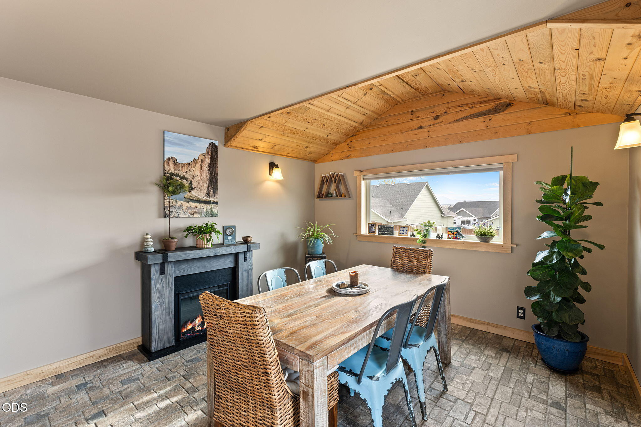 Dining room with a vaulted wood ceiling, rustic table, fireplace, and window overlooking neighboring homes.