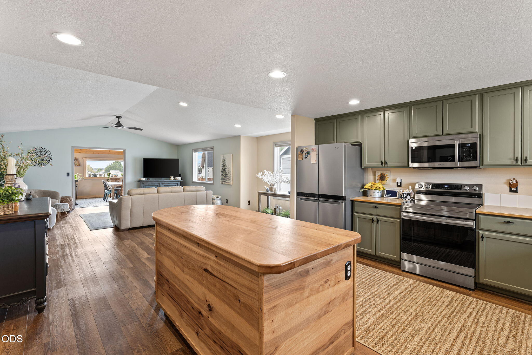 Open-plan kitchen with a large wooden island, sage cabinets, and living room beyond.